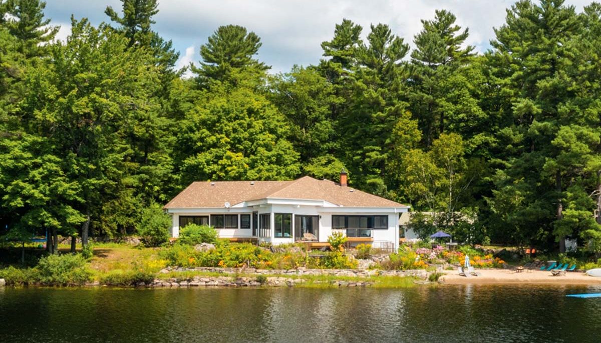 White cottage with a brown roof surrounded by trees with a lake in front.