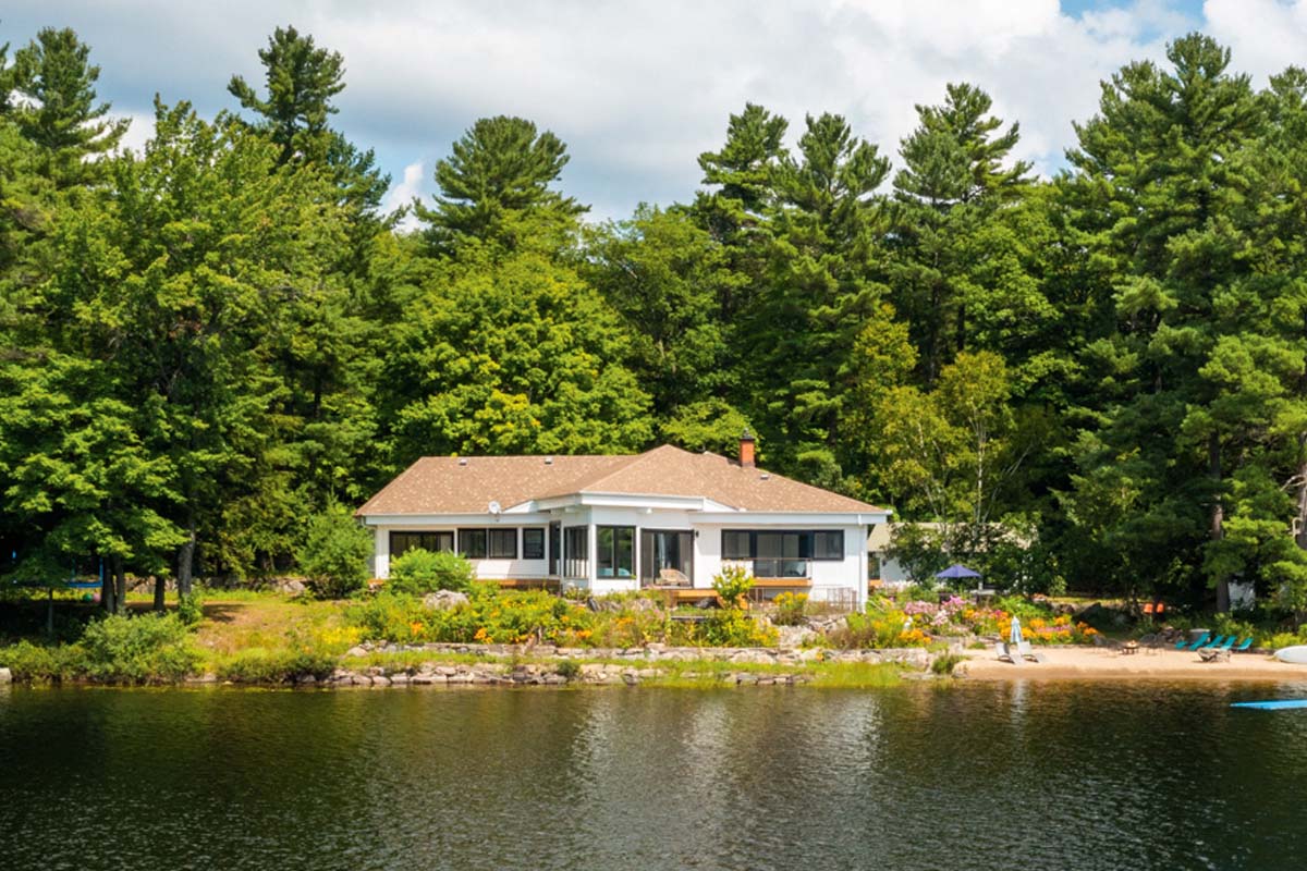 White cottage with a brown roof surrounded by trees with a lake in front.