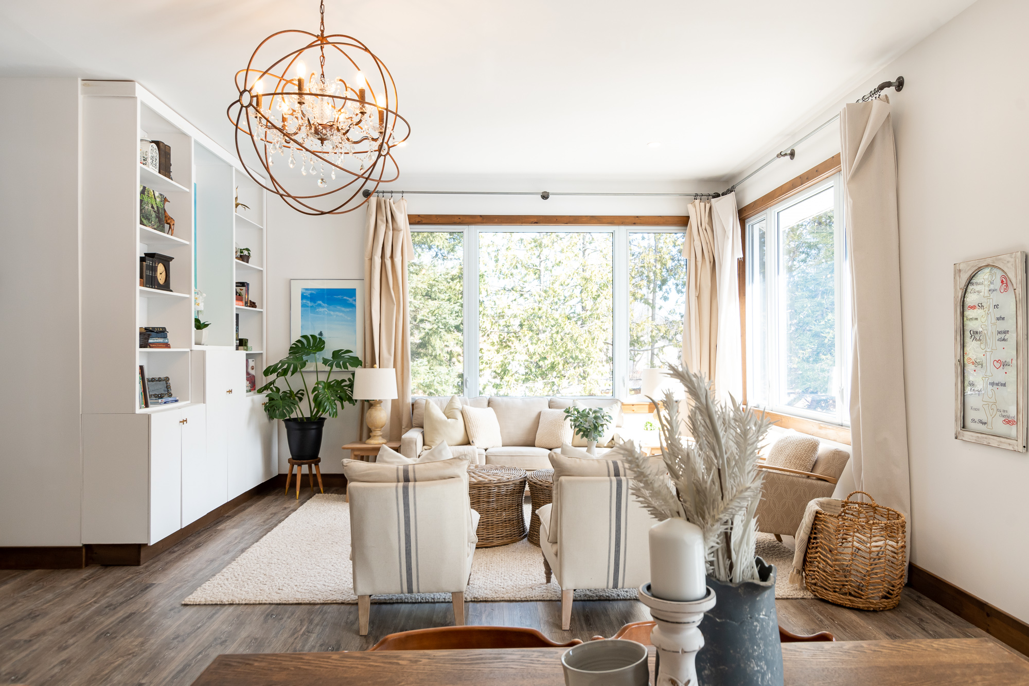 Bright living area surrounded by windows. White chairs and a white couch face each other, to the left is a white bookshelf