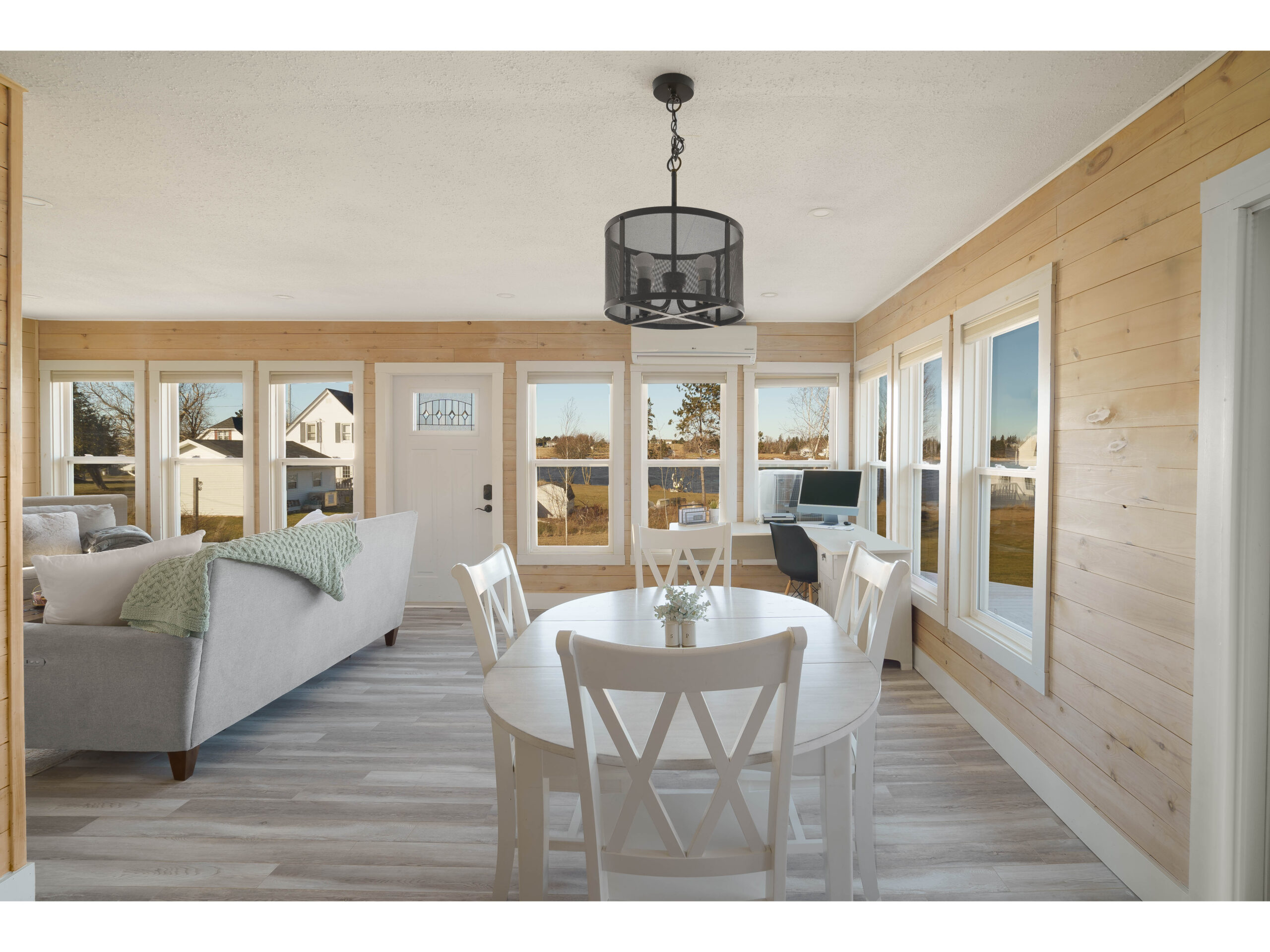 An open-concept dining and living space just inside a front door. Lots of windows let in natural light, and light wood panelling covers the walls.