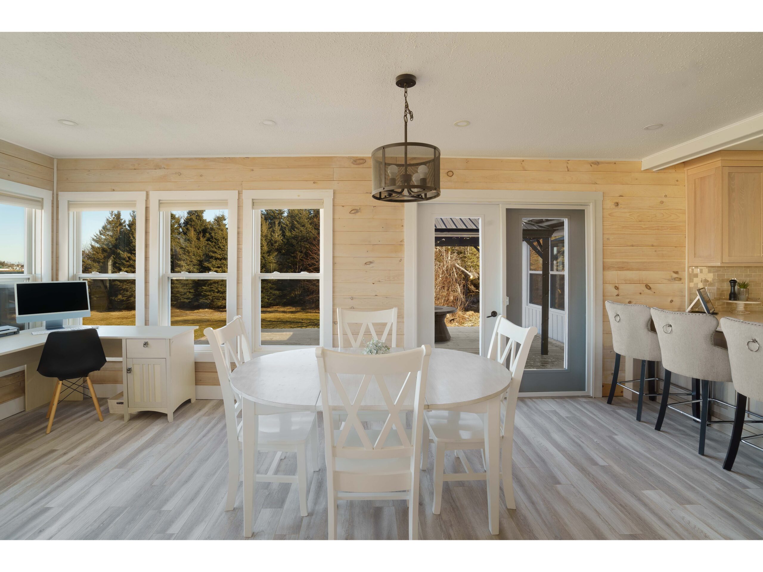 An open-concept dining area. Double doors open out to a big outdoor deck. Lots of windows let in natural light, and light wood panelling covers the walls.