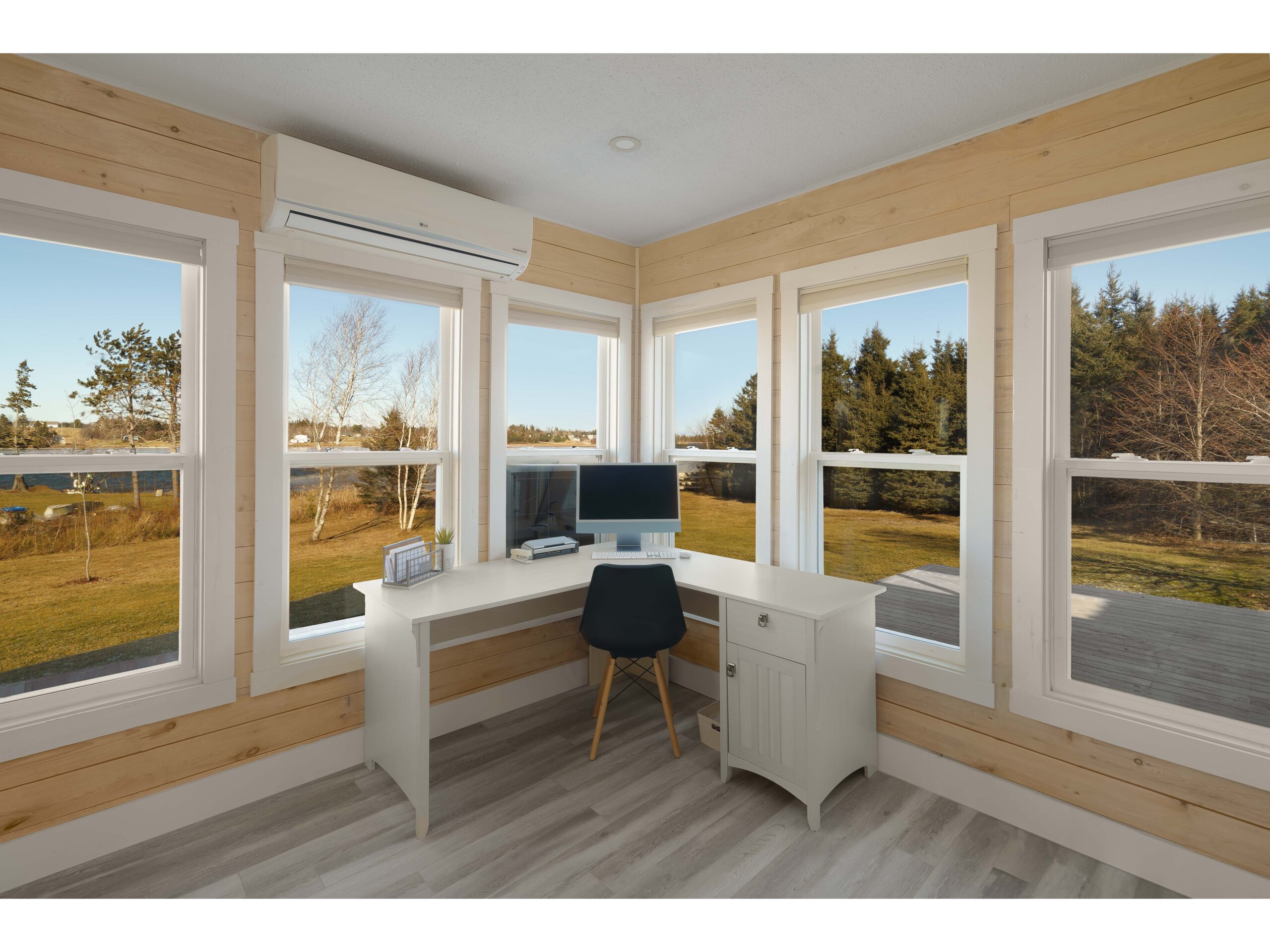 A minimal white corner desk, with a black chair and a computer monitor, sits nestled in the corner of wood-panelled walls with lots of big windows.