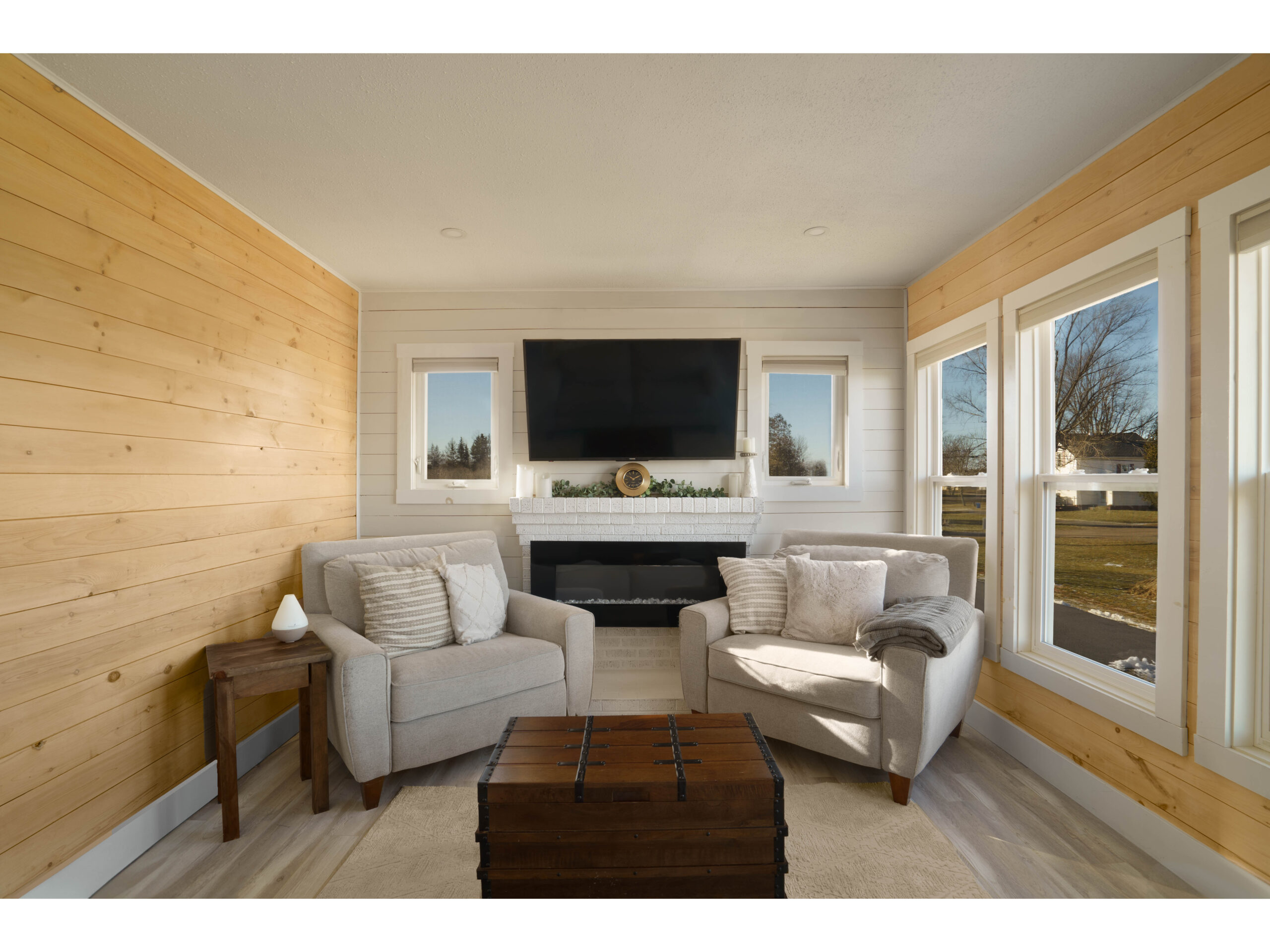 A bright but cozy living space with big windows, two chairs, a television, and wood-panelled walls.