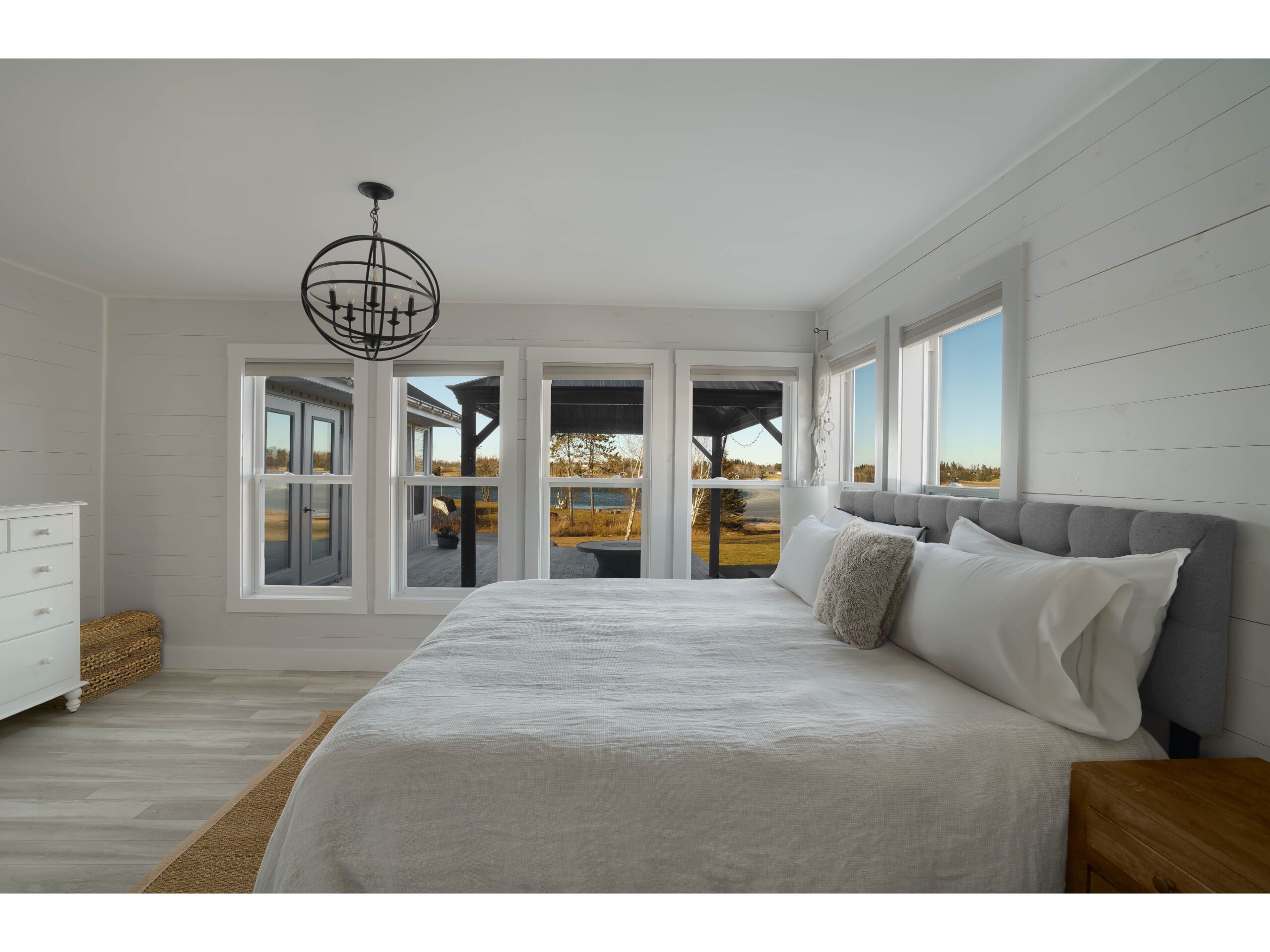 A large primary bedroom with a big bed, a modern light fixture, a dresser, and lots of windows, looking out toward the water.