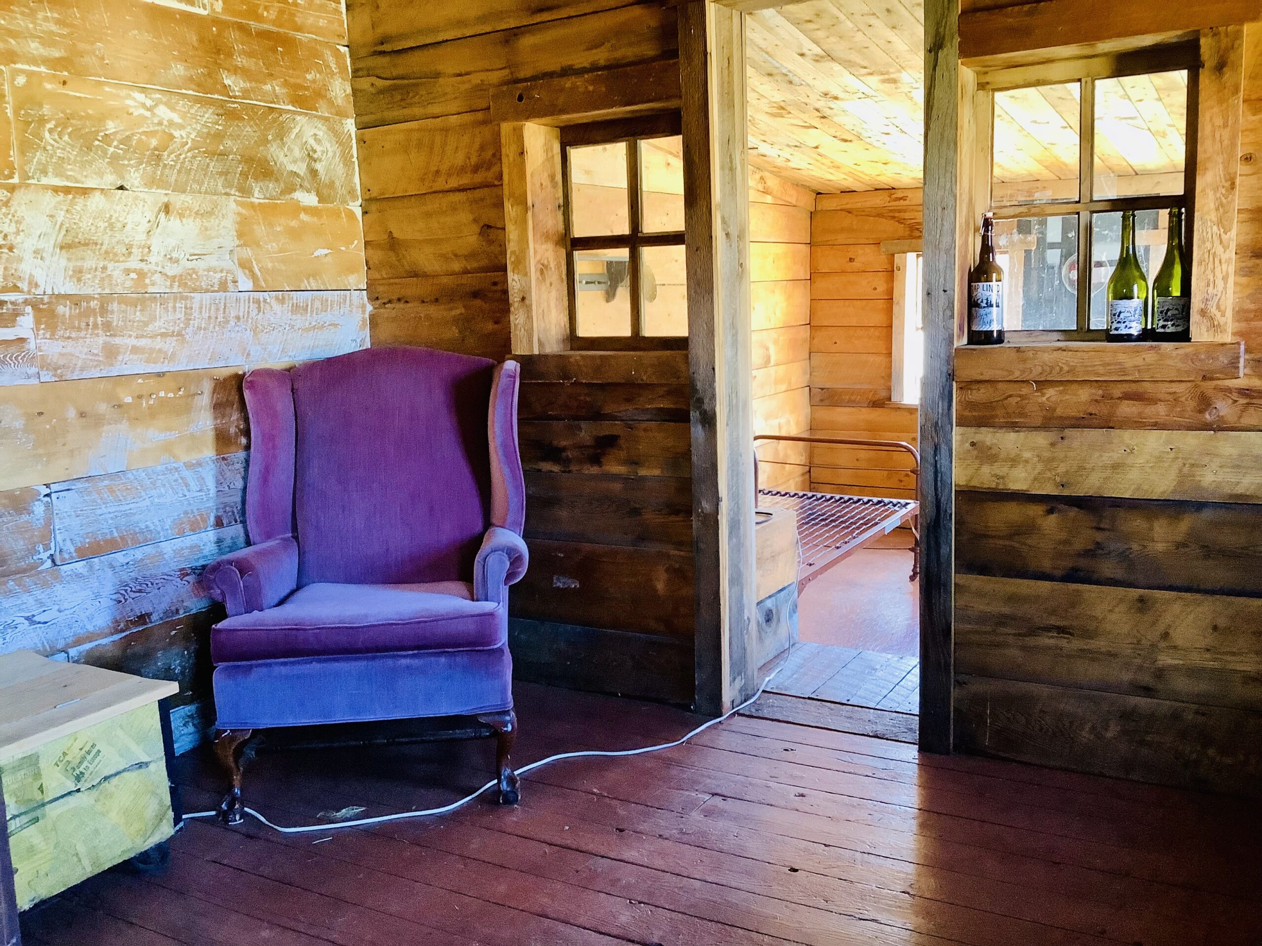 Interior of a small, rustic wood bunkie. A pink plush chair sits in the corner of a room, and through a doorframe there is another room with an empty metal bed frame.
