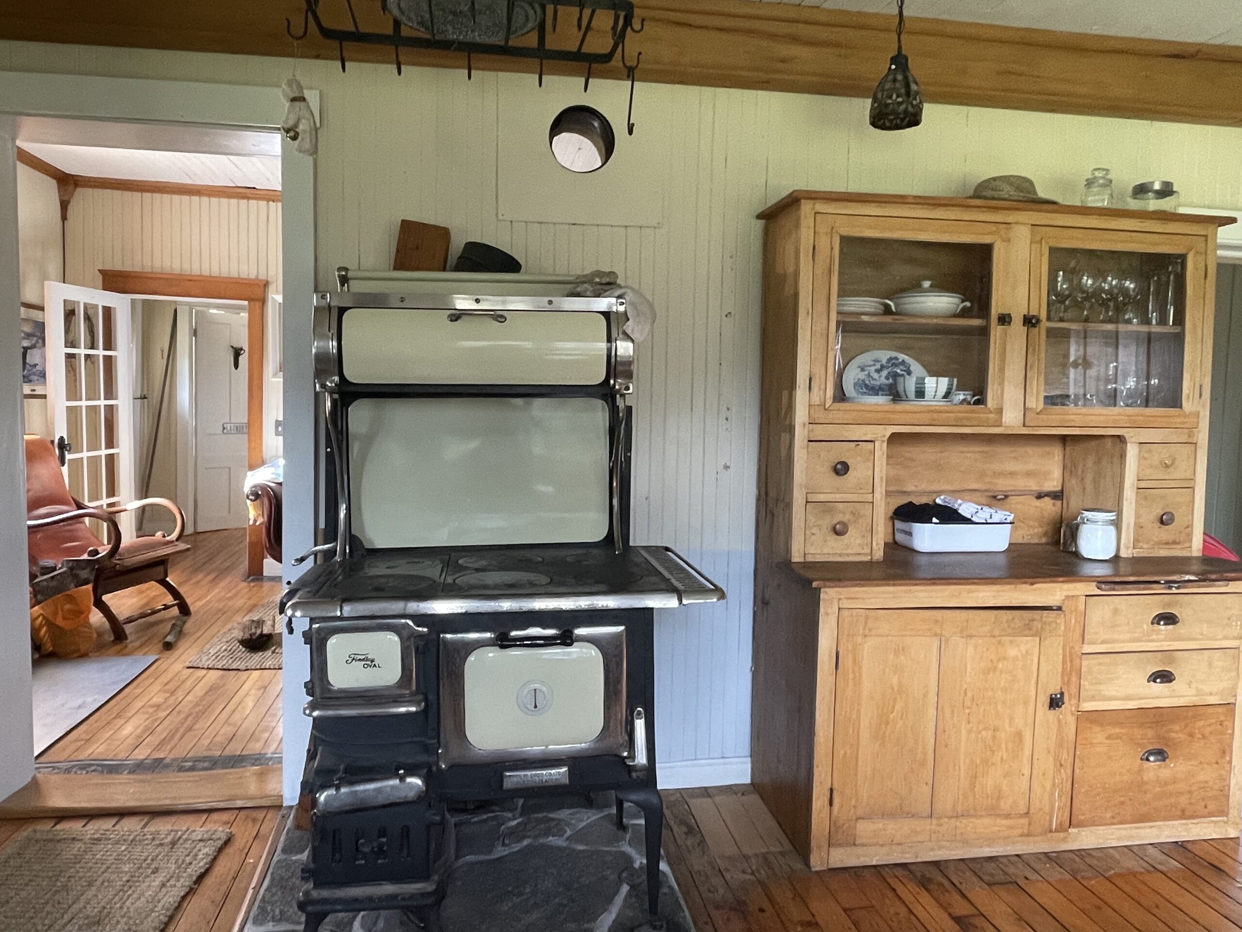 An old stove and a tall wooden cabinet with a desk surface and glass doors, against the wall in a kitchen.