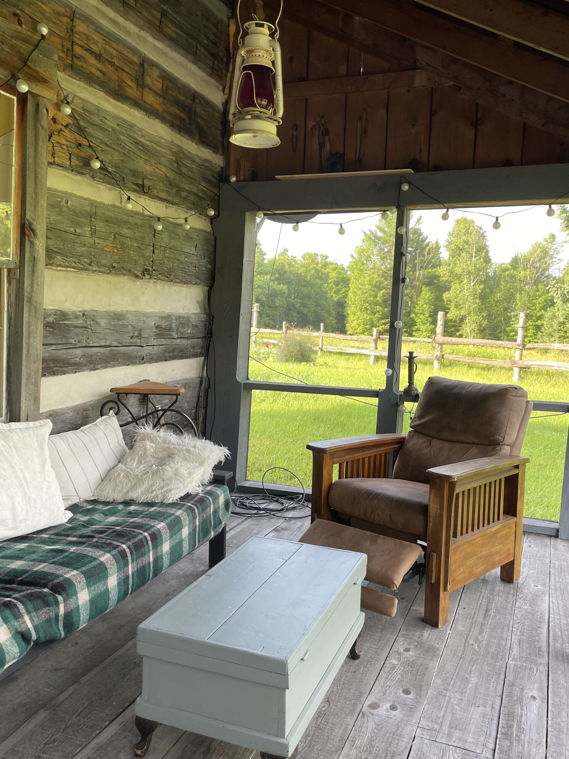 A couch, a coffee table, and a chair on a screened-in porch. Lights are strung overhead.