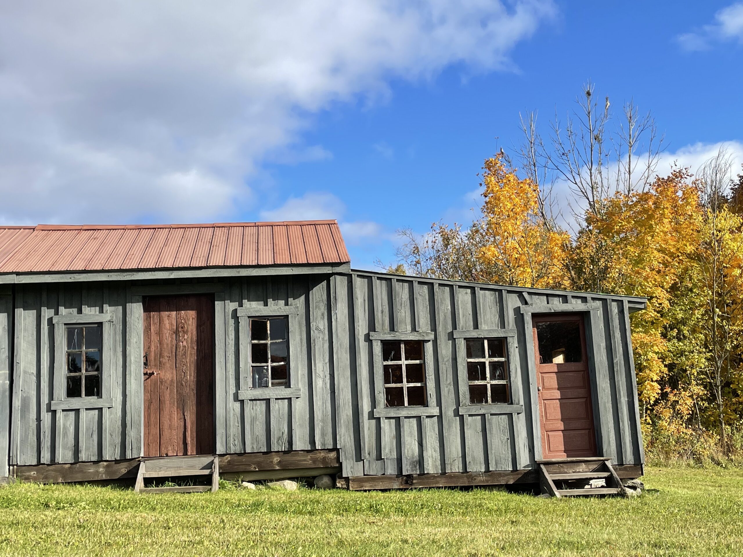 The front of a small wooden bunkie with gray wood exterior, two red doors, and a red roof.