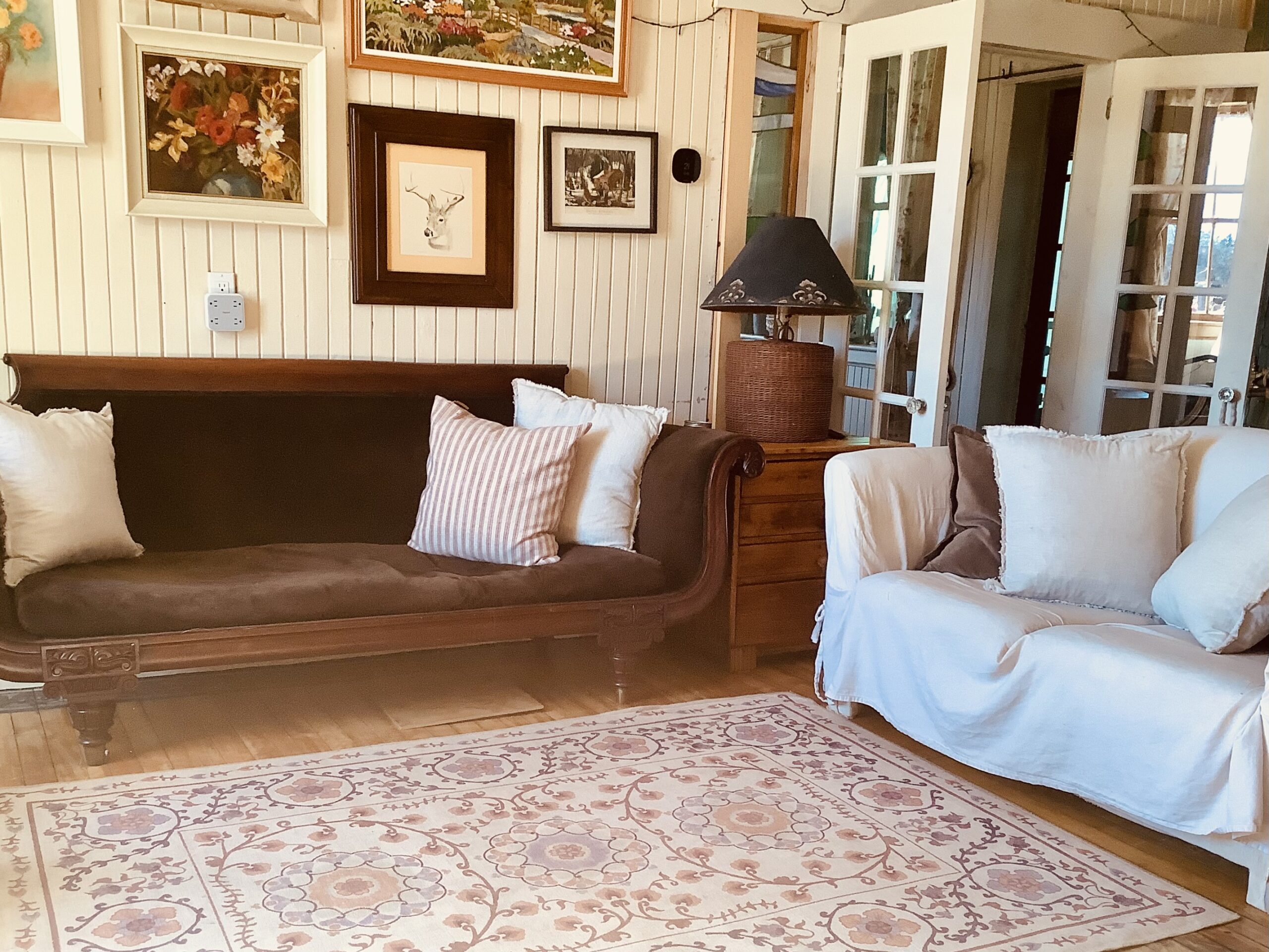A small, bright living area with a vintage brown couch, a white sofa, and an ornate floral rug over hardwood flooring. A gallery wall of framed art hangs above the couch.