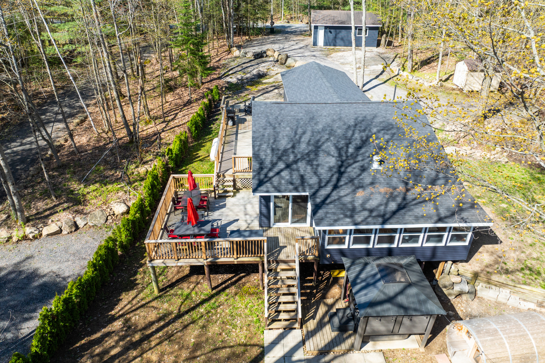 Overhead view of a large cottage with navy blue exterior siding, lots of windows, and a large two-level wooden deck with an outdoor dining area. Across a big driveway is a small detached garage.
