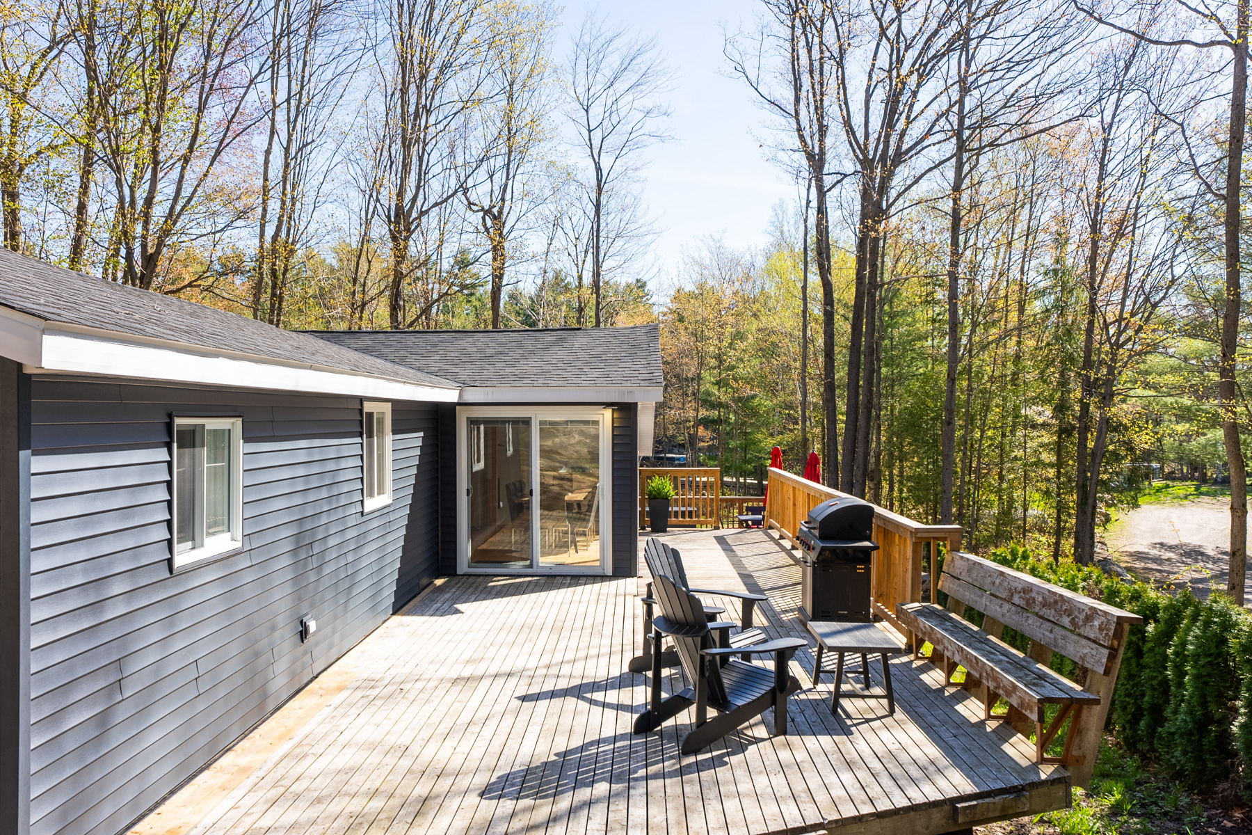 A big wooden deck off a cottage with navy blue exterior siding. Muskoka chairs and a bench sit next to a barbecue. Sliding glass doors lead into the cottage.