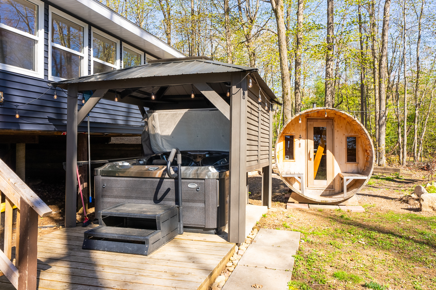 A hot tub underneath a privacy structure. A wooden barrel sauna sits behind the hot tub area.