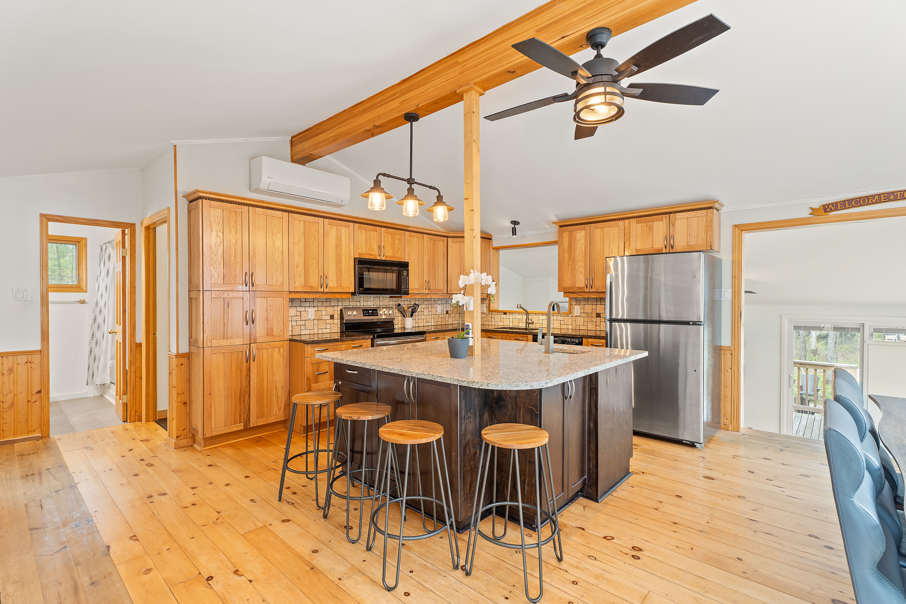 A large, open-concept kitchen with warm wood hardwood flooring, a huge island with a granite countertop and barstool seating, wood cabinetry, and a vaulted ceiling with a wooden beam across the top.