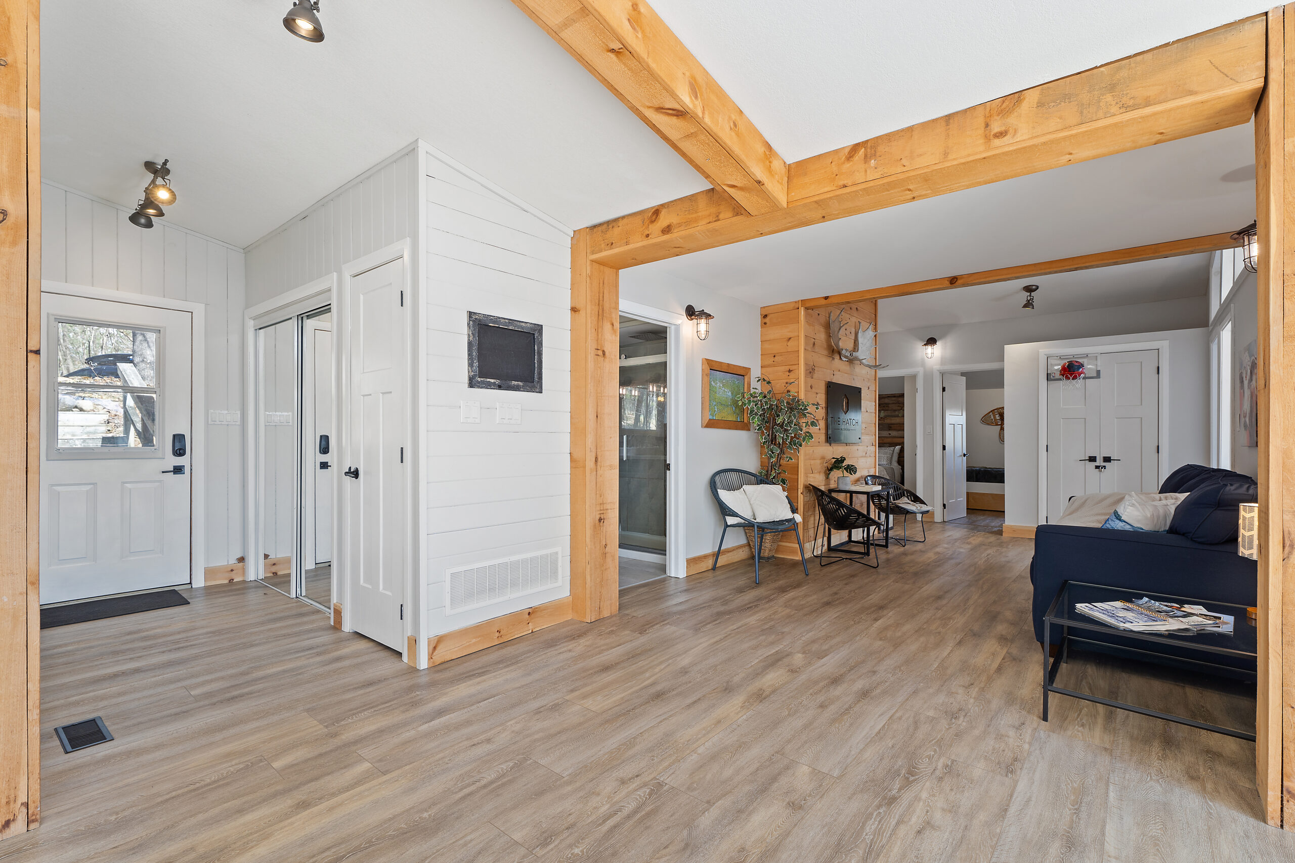 Entrance to the cottage with a white door on the left and open space to rest of the cottage on the right. Light brown floors and red-brown wood beams line the room