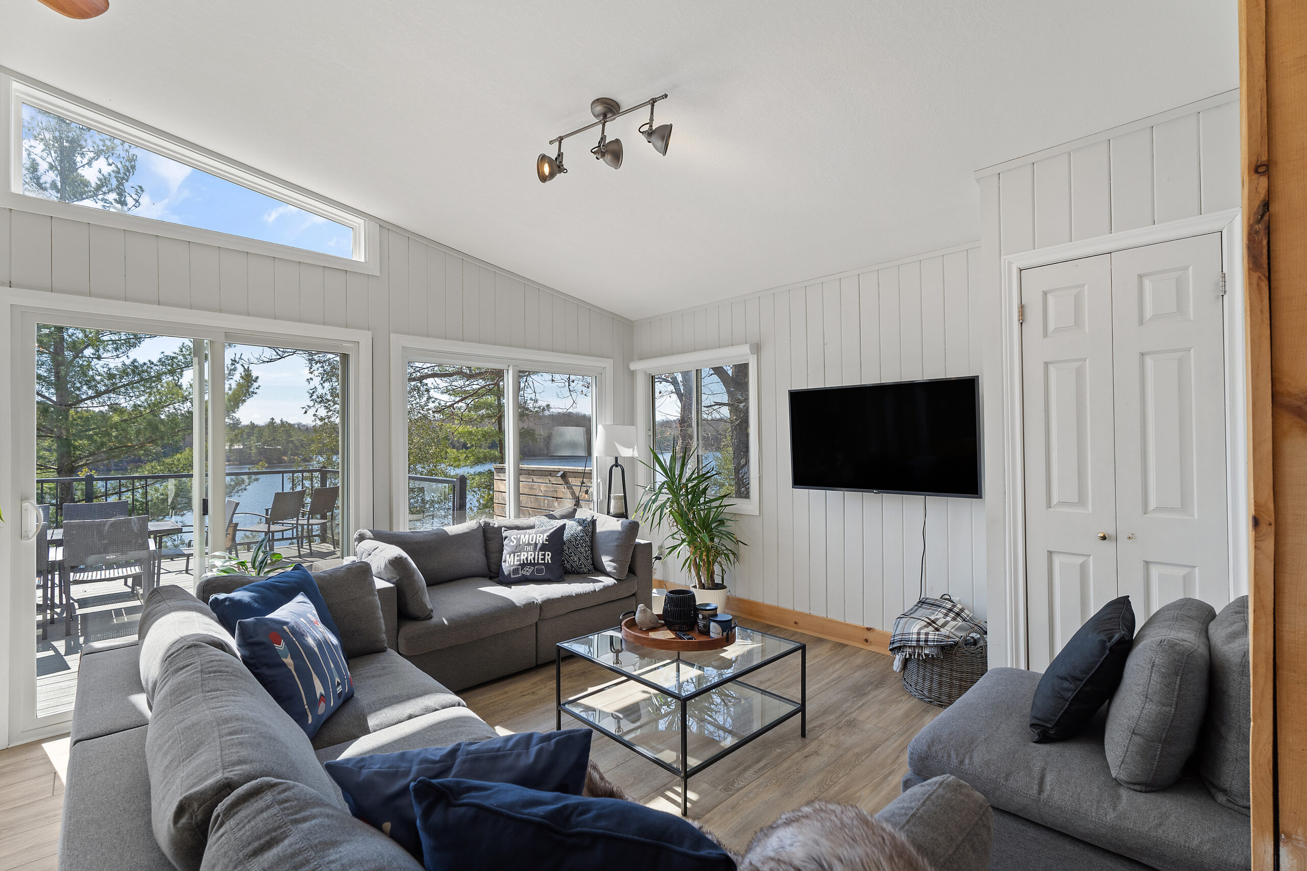 A living area with grey couches and chairs surround a glass table, facing a white wall with a flatscreen TV mounted