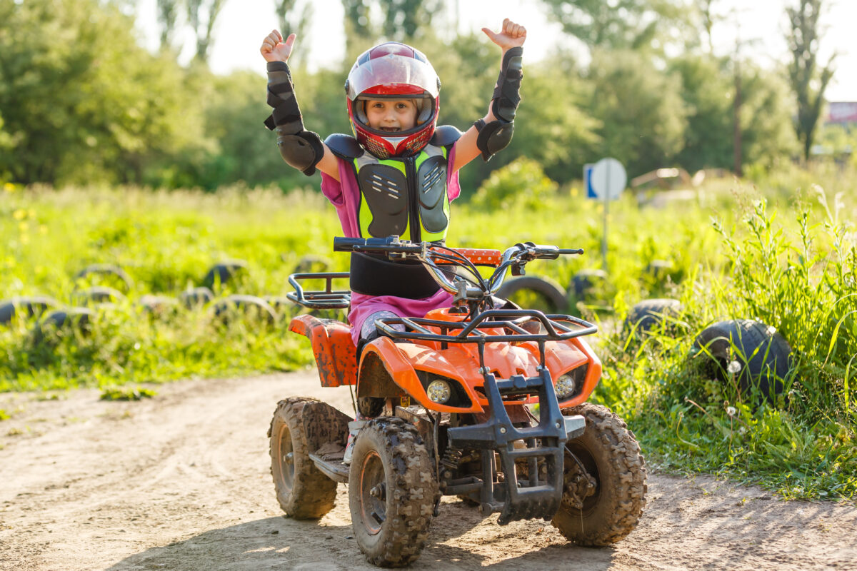 A young girl wearing a helmet and protective gear gives a double-thumbs-up while sitting on an ATV