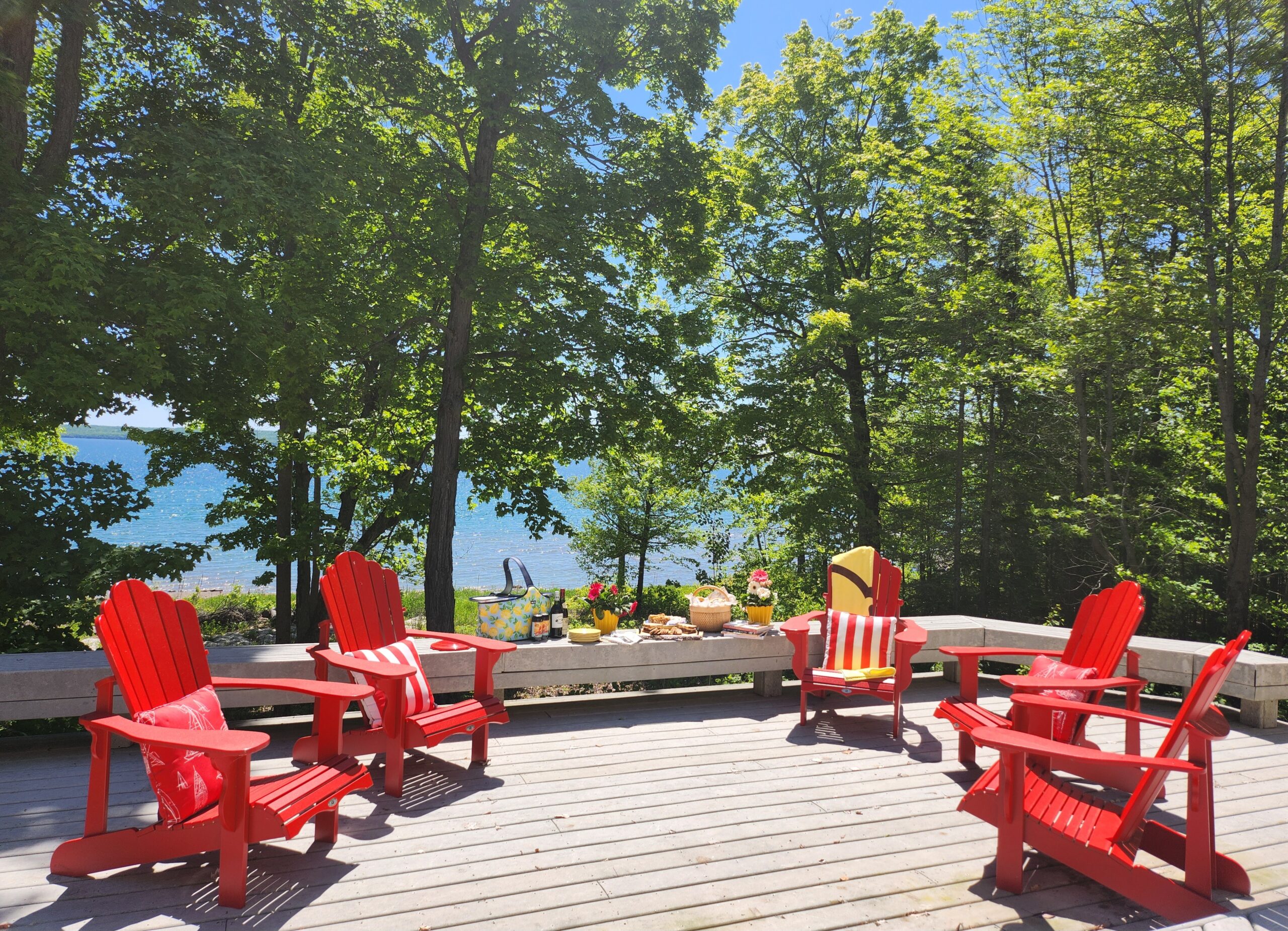 Red Muskoka chairs face each other. They sit at the edge of the deck with lush green trees behind and lake views.