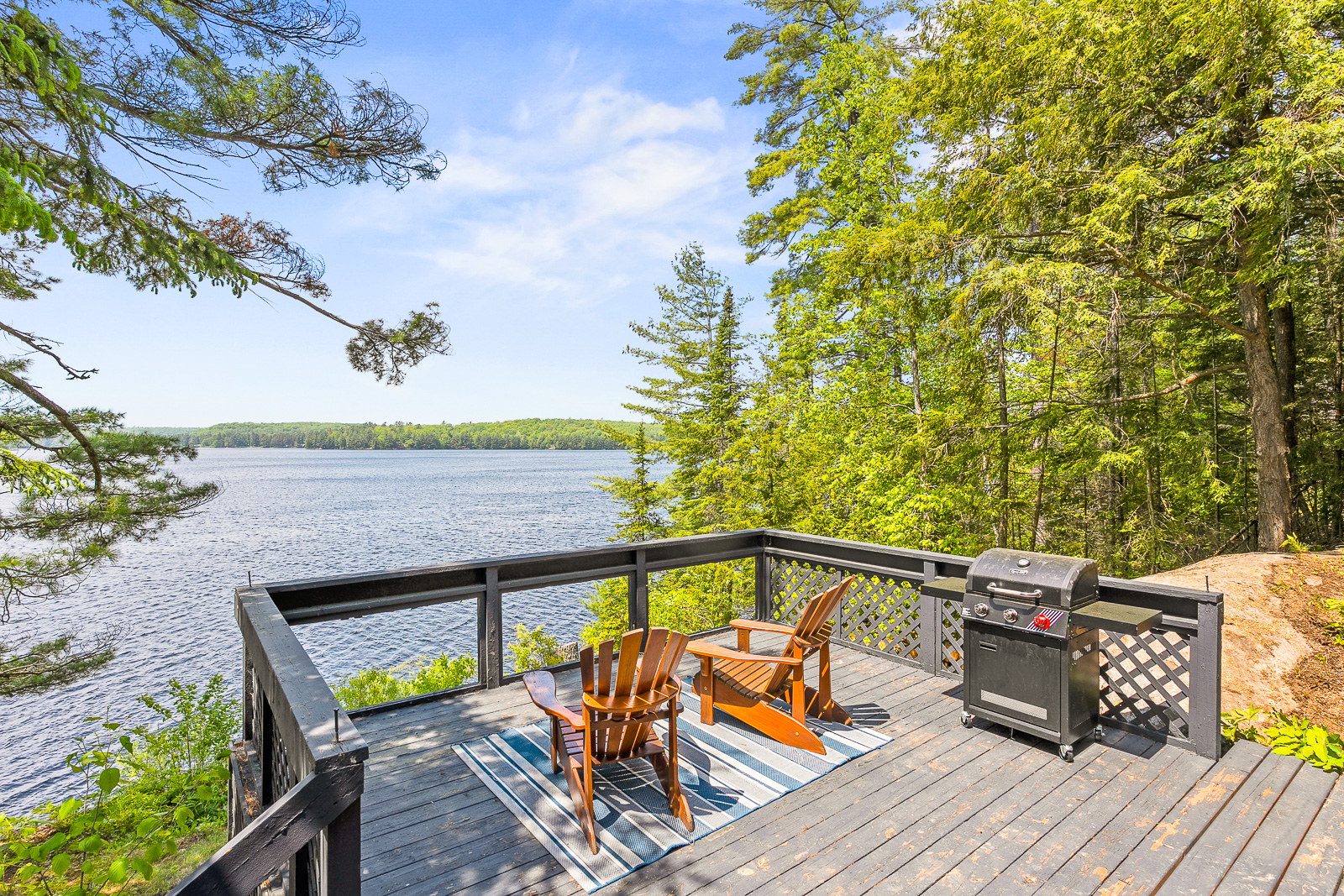 A grey deck with glass rails overlooks the lake. Two brown Muskoka chairs on a blue carpet face the lake