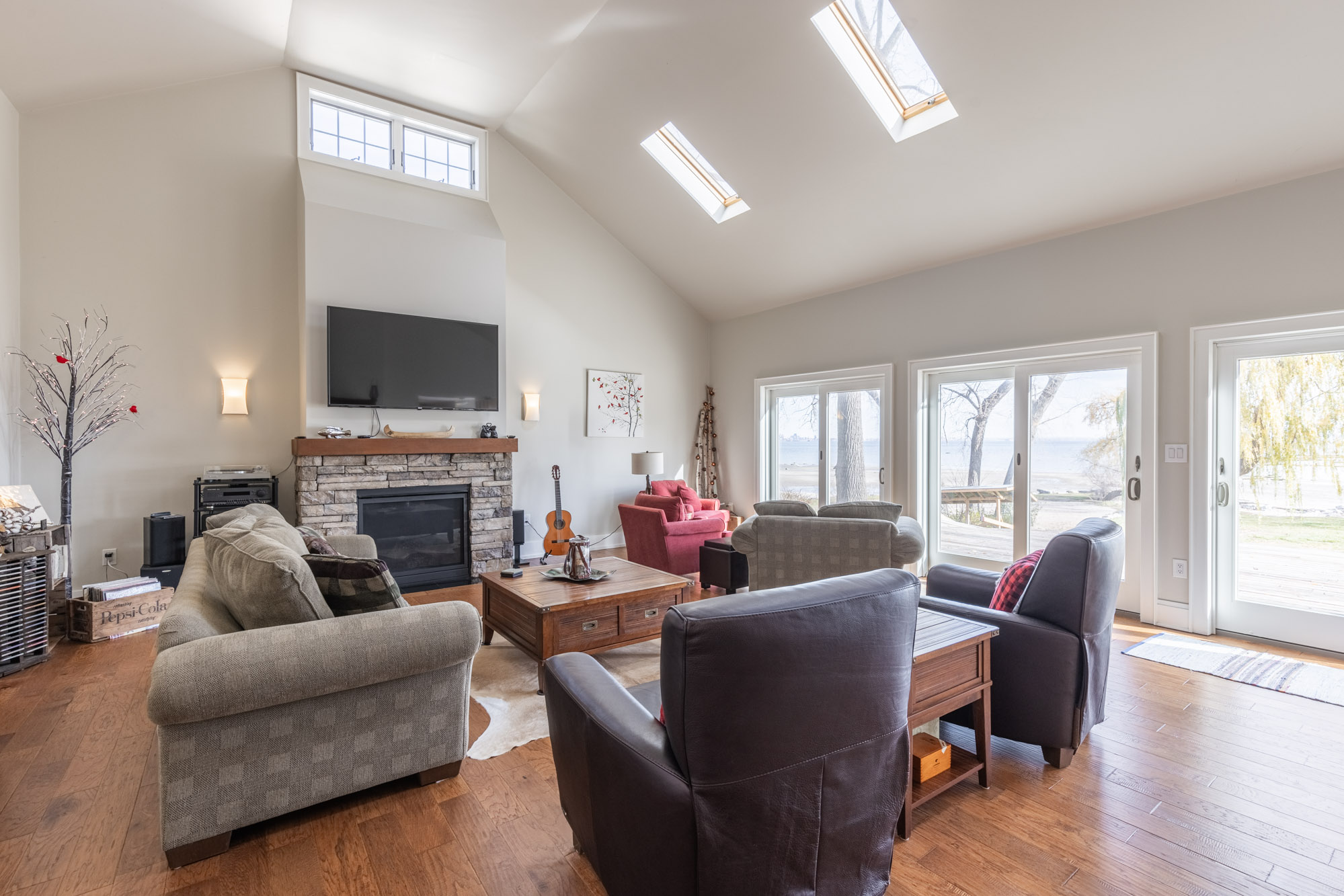 A bright family room with white, vaulted ceilings with skylights. Couches and chairs face each other, a TV hangs above the fireplace