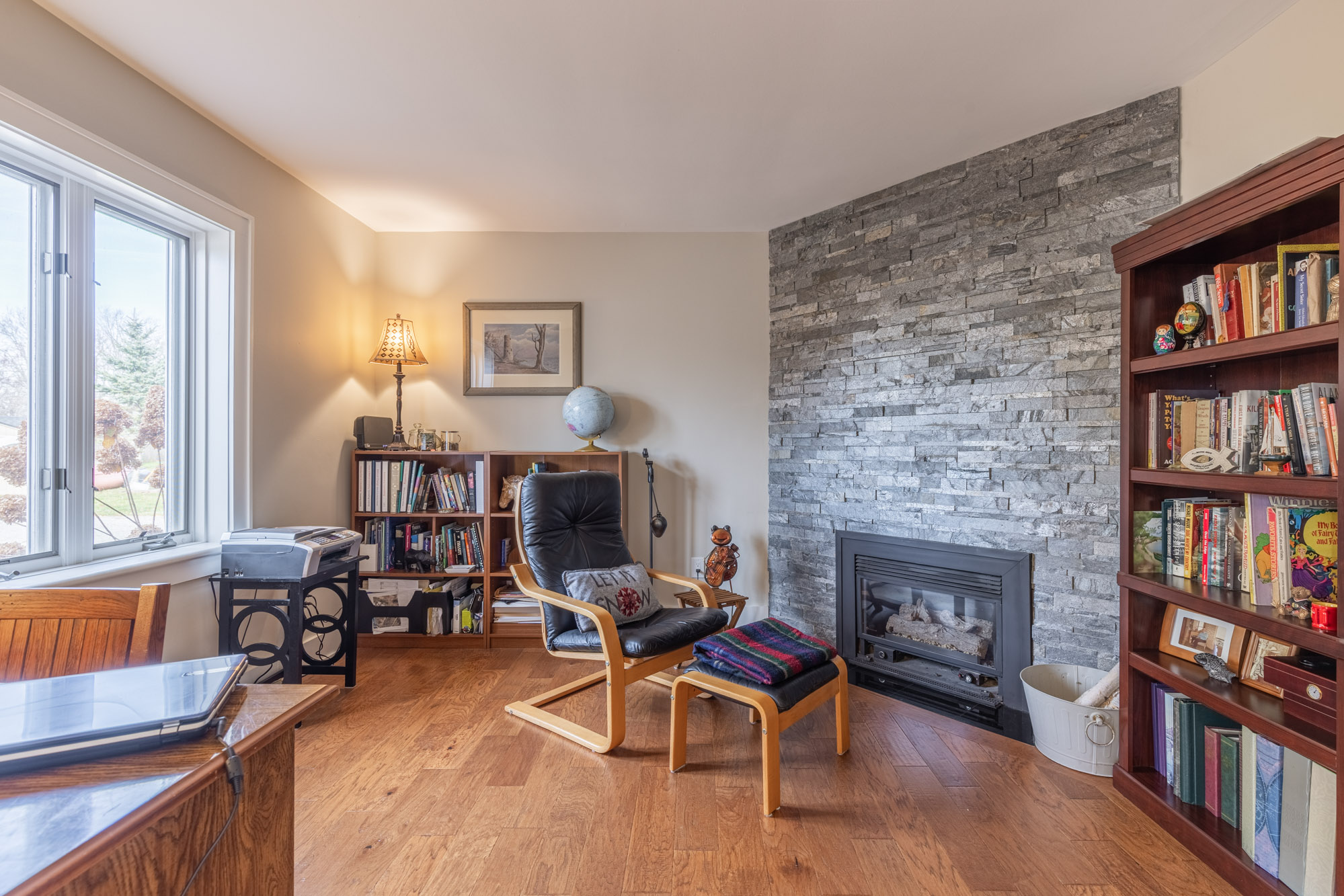 Brown hardwood floods line the room, a small faces a large book shelf. Part of the wall is covered in grey stone with a small black fireplace at the bottom