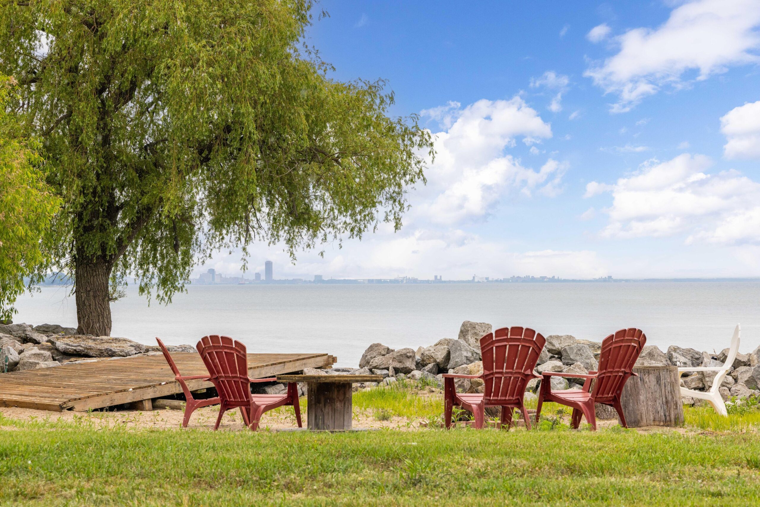 Red Muskoka chairs around a fire pit with the lake and a large tree in the background