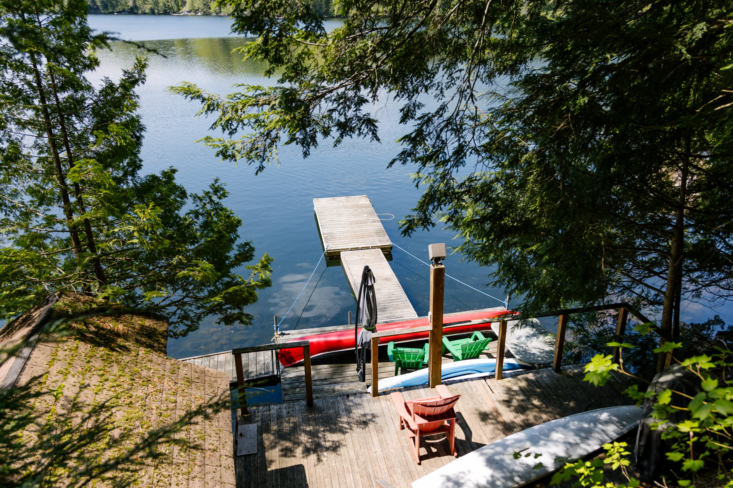 A dock in blue water. On the shore is a red canoe and green Muskoka chairs