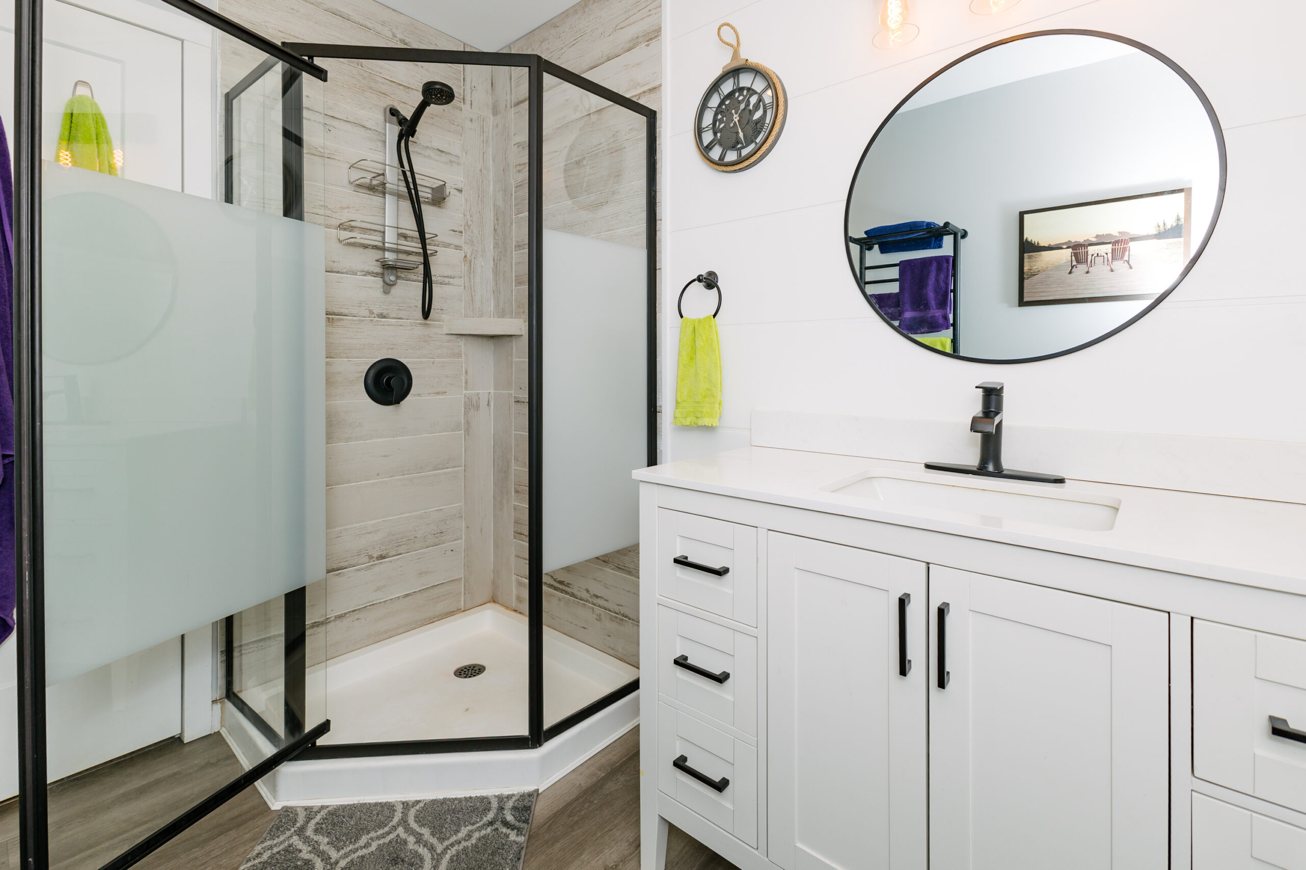 A glass shower with black trim is open to reveal panelled walls. To the right, a white cabinet with a round mirror above