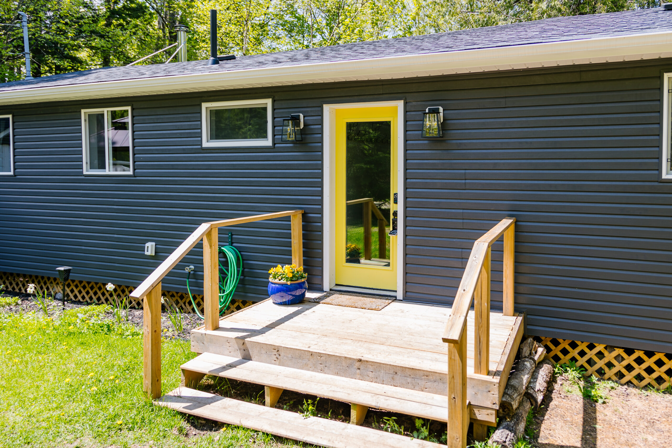 A bright yellow glass front door on a grey-blue panelled bungalow. Wood steps with rails lead up to the door