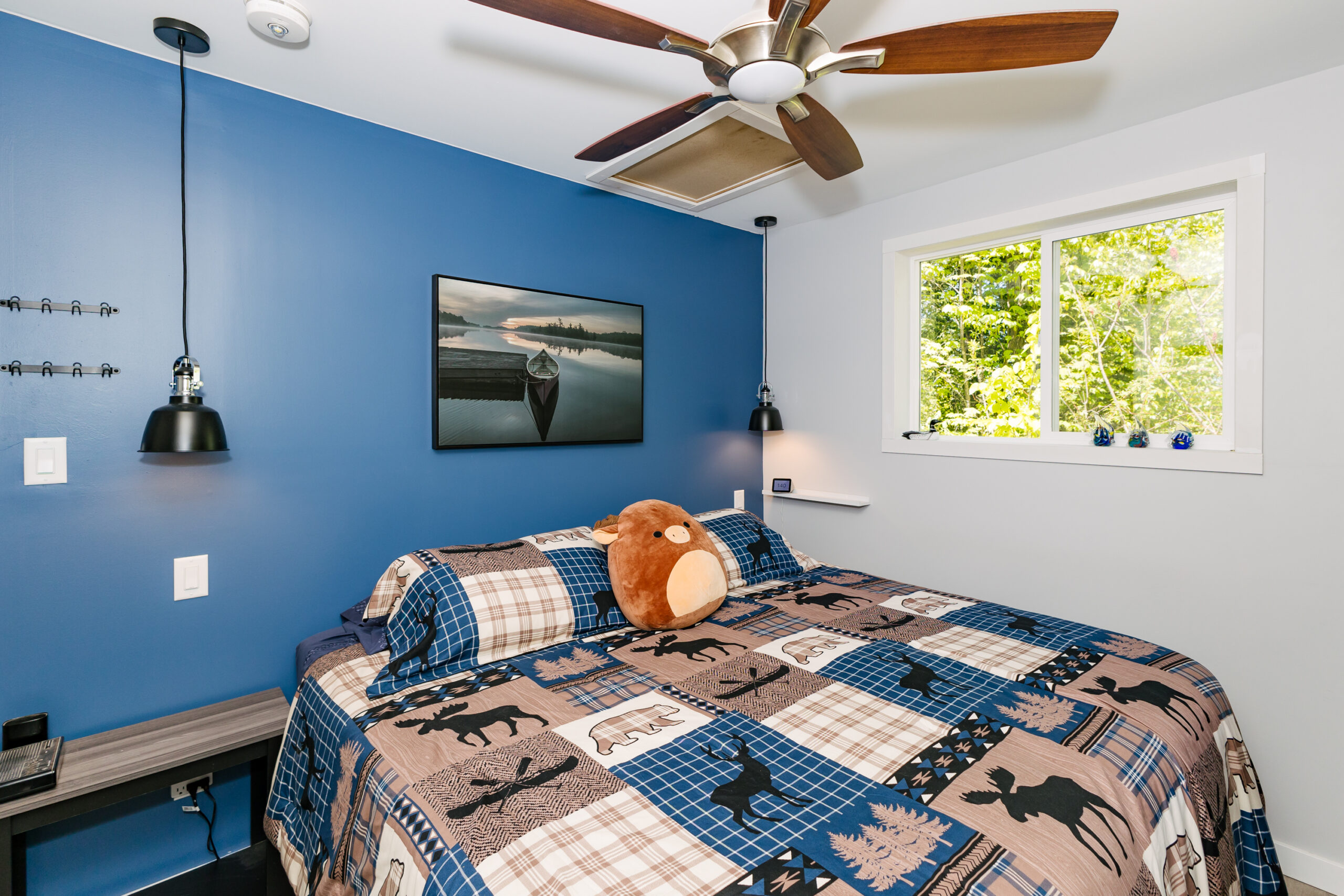 A bed with a blue and brown patterned bedspread sits against a blue wall. A brown ceiling fan above.