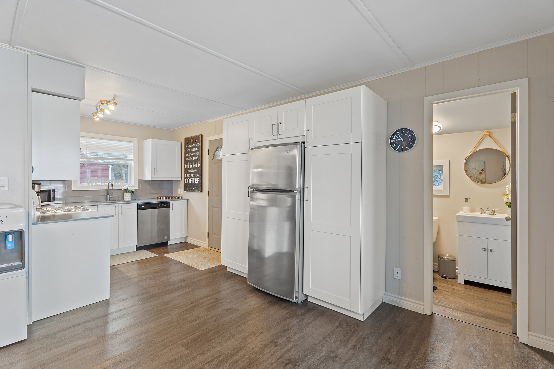 A bright, open space with a kitchen just off a door to outside. The kitchen is small, with stainless steel appliances and lots of white cupboards. A doorway to the right of the kitchen holds a bathroom.
