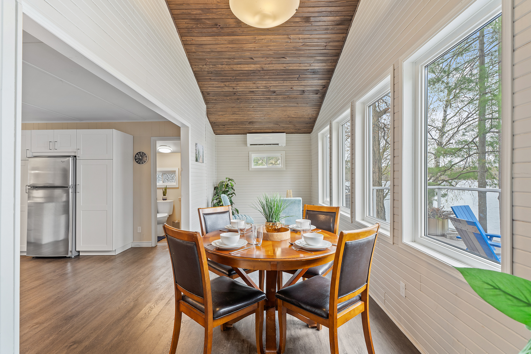 A small dining table and chairs in front of big windows, with a sloping wood-paneled ceiling overhead.