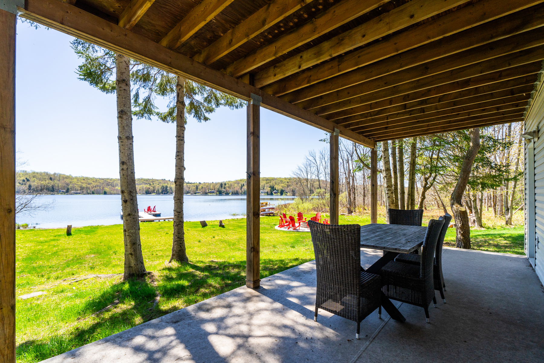 Side angle of a covered lower-level deck with a dining table. In the background, the grassy backyard and lake