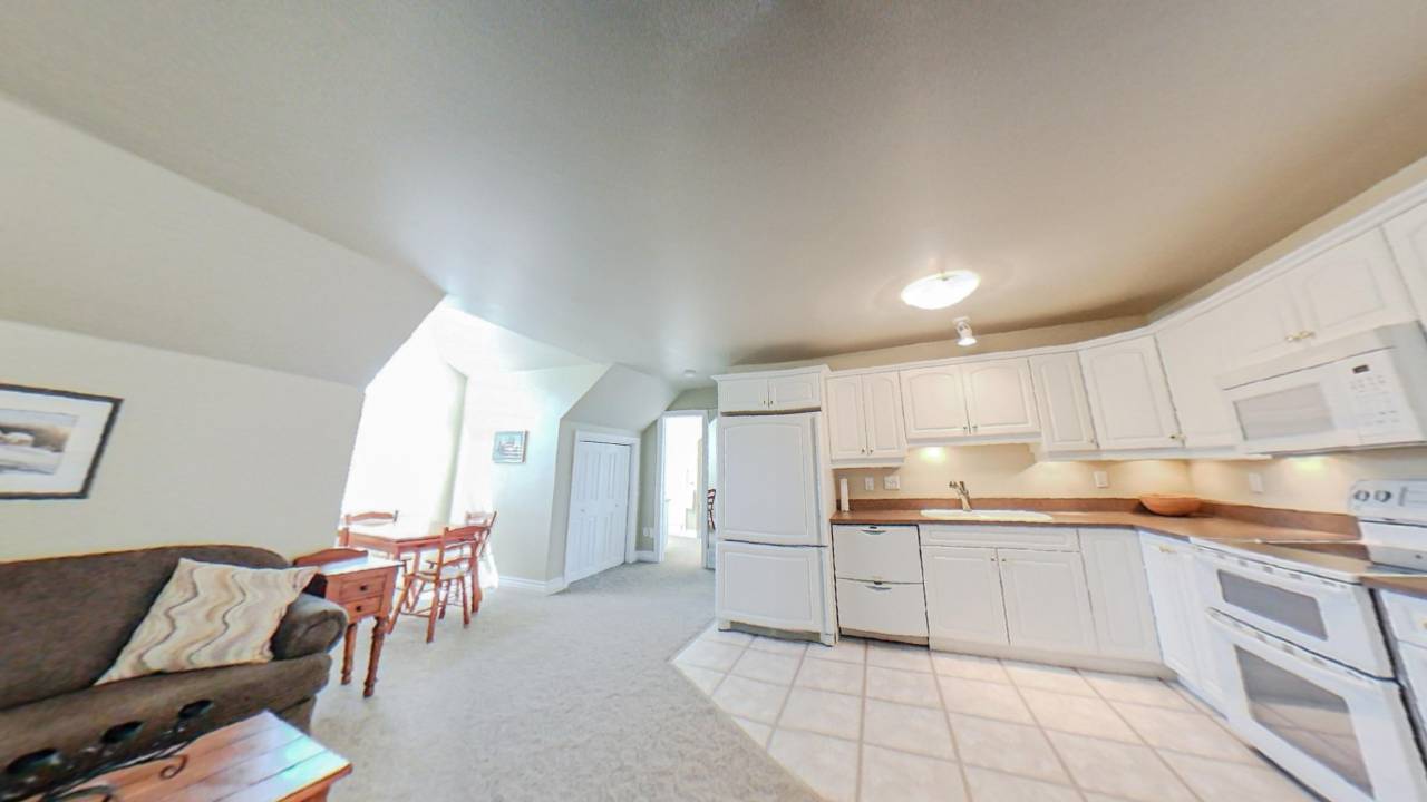 A white room with a small wood dining set next to a window. To the right, a white kitchen