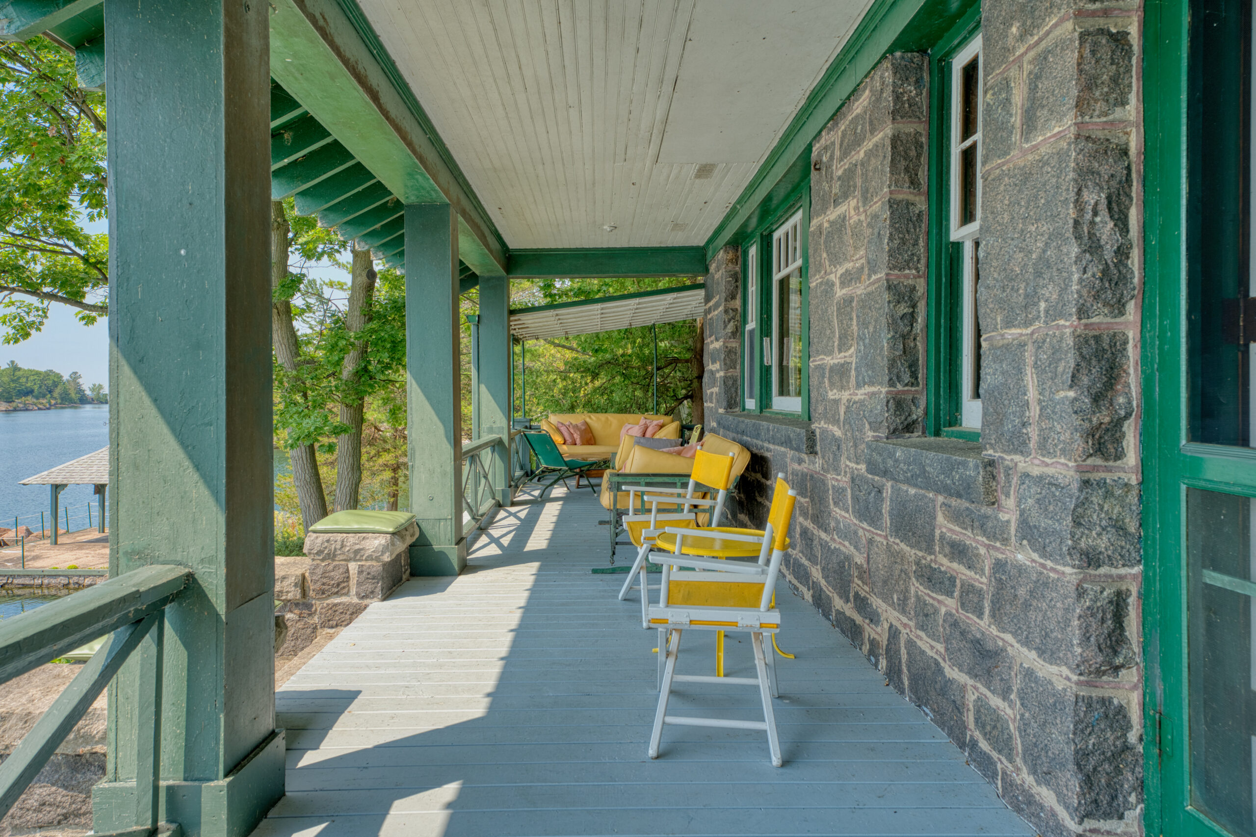 A covered porch in front of the stone house. White chairs with yellow cushions line the porch