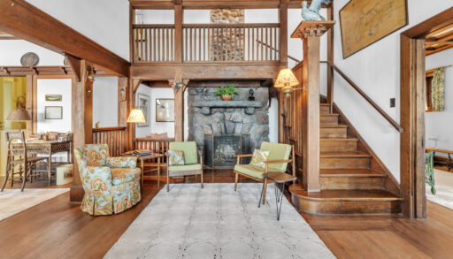 A grand foyer with wood fixtures. A large stone fireplace in the centre is framed by two light green chairs sitting on a large cream carpet. To the right, a wood staircase leads up to the second floor