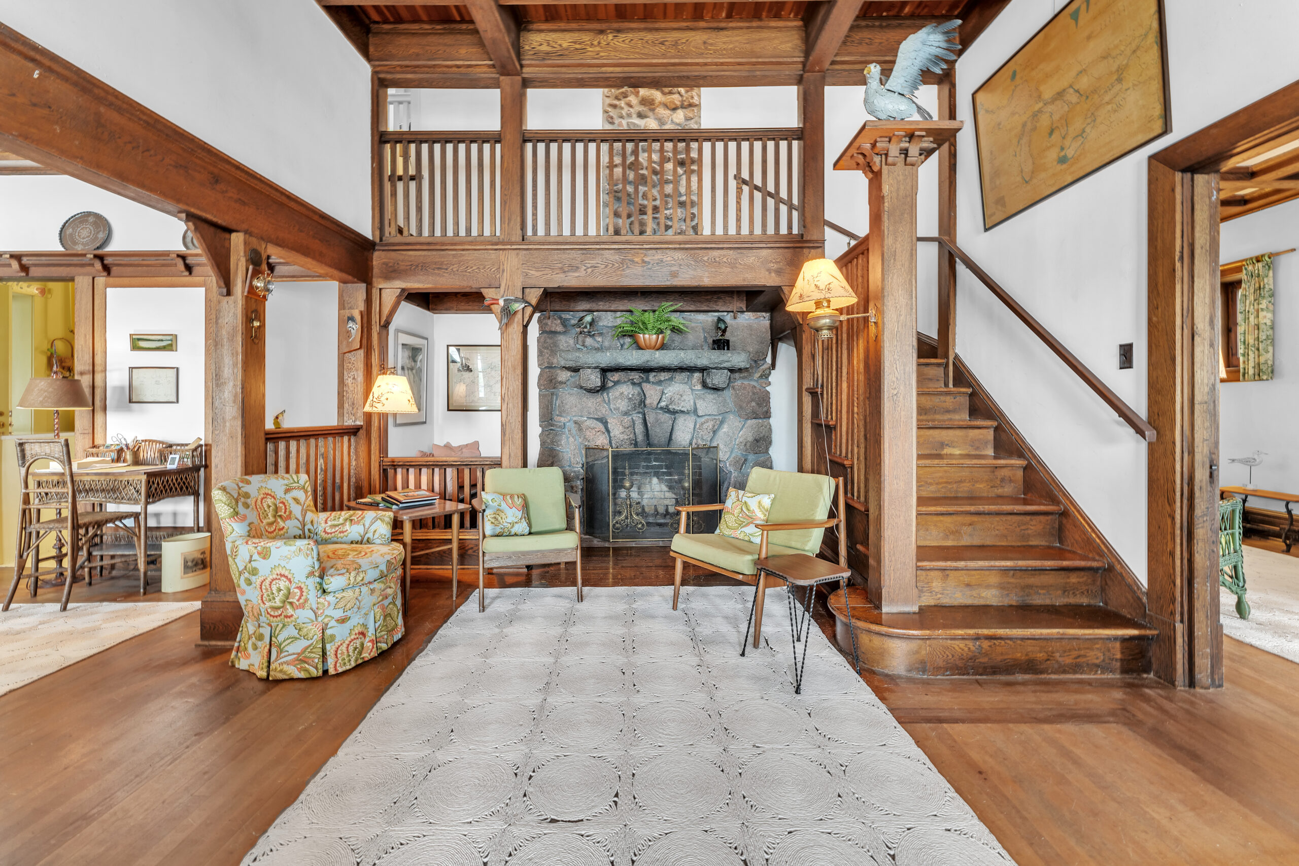 A grand foyer with wood fixtures. A large stone fireplace in the centre is framed by two light green chairs sitting on a large cream carpet. To the right, a wood staircase leads up to the second floor