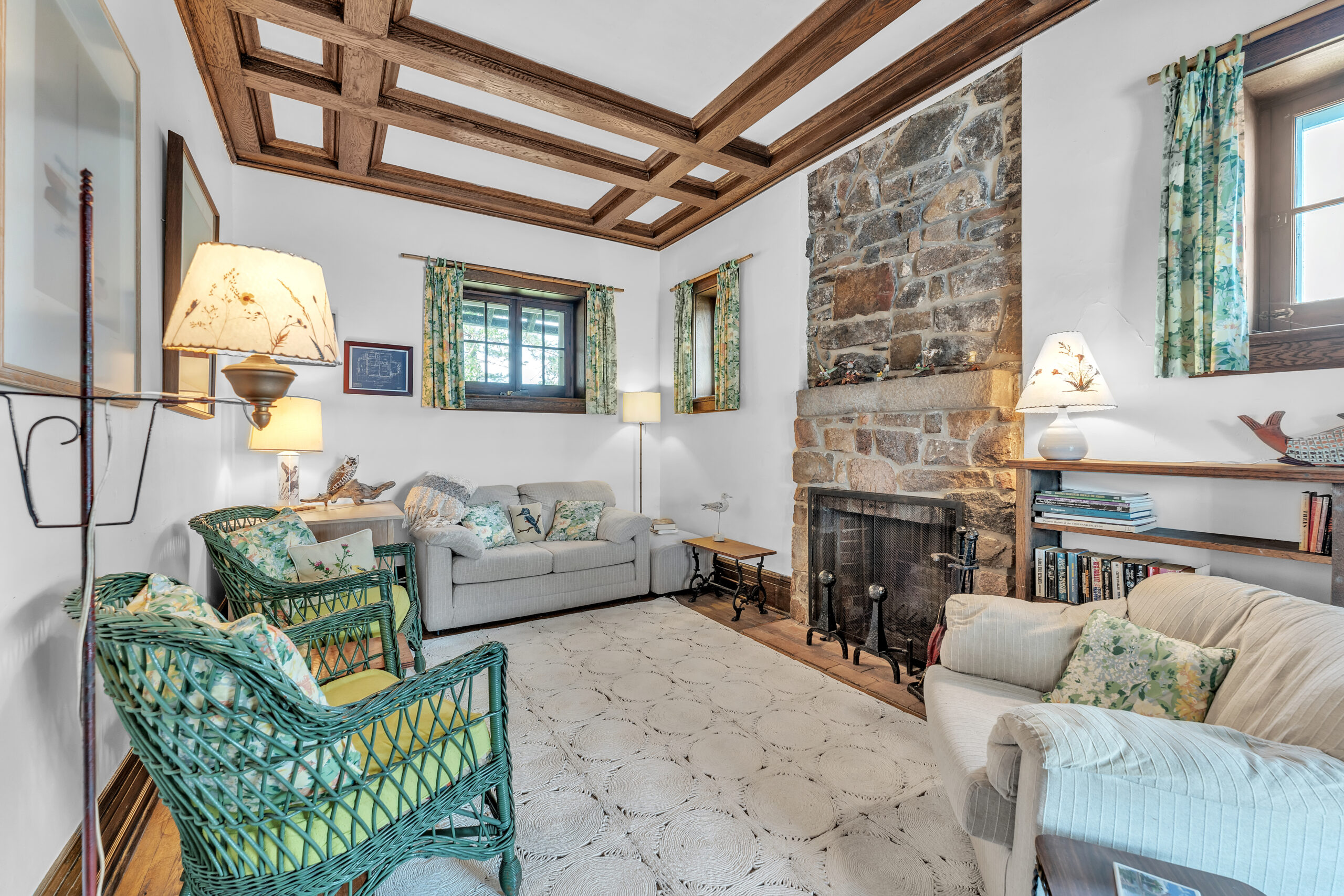 A white couch perpendicular to two green wicker chairs and a large stone fireplace