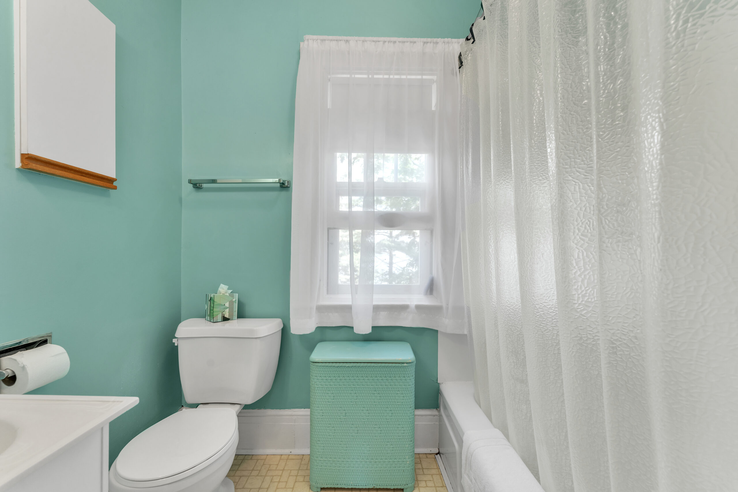 A teal bathroom with a white toilet, bright window with a sheer curtain, and a bathtub