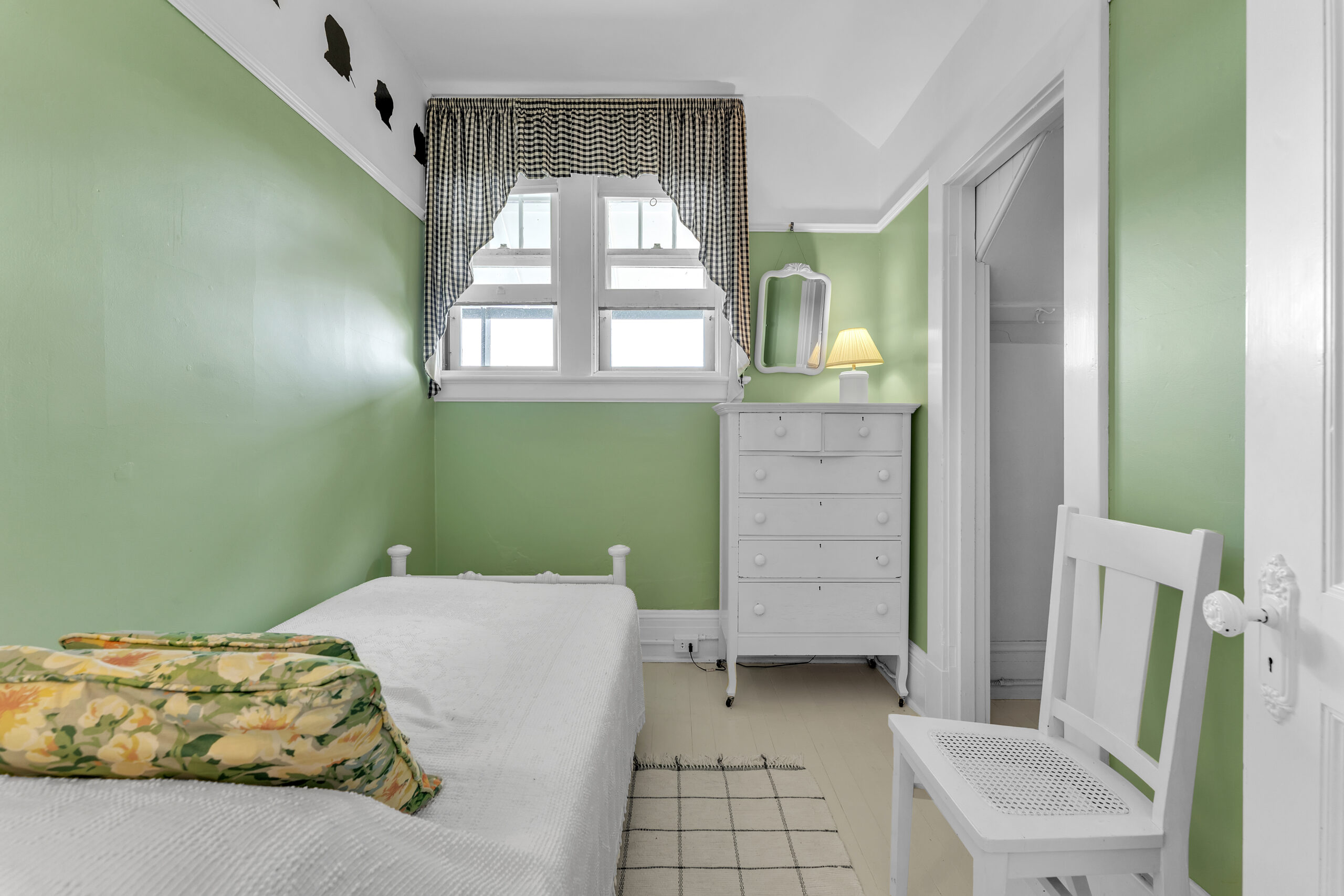 A white twin bed in a painted green room faces a high window