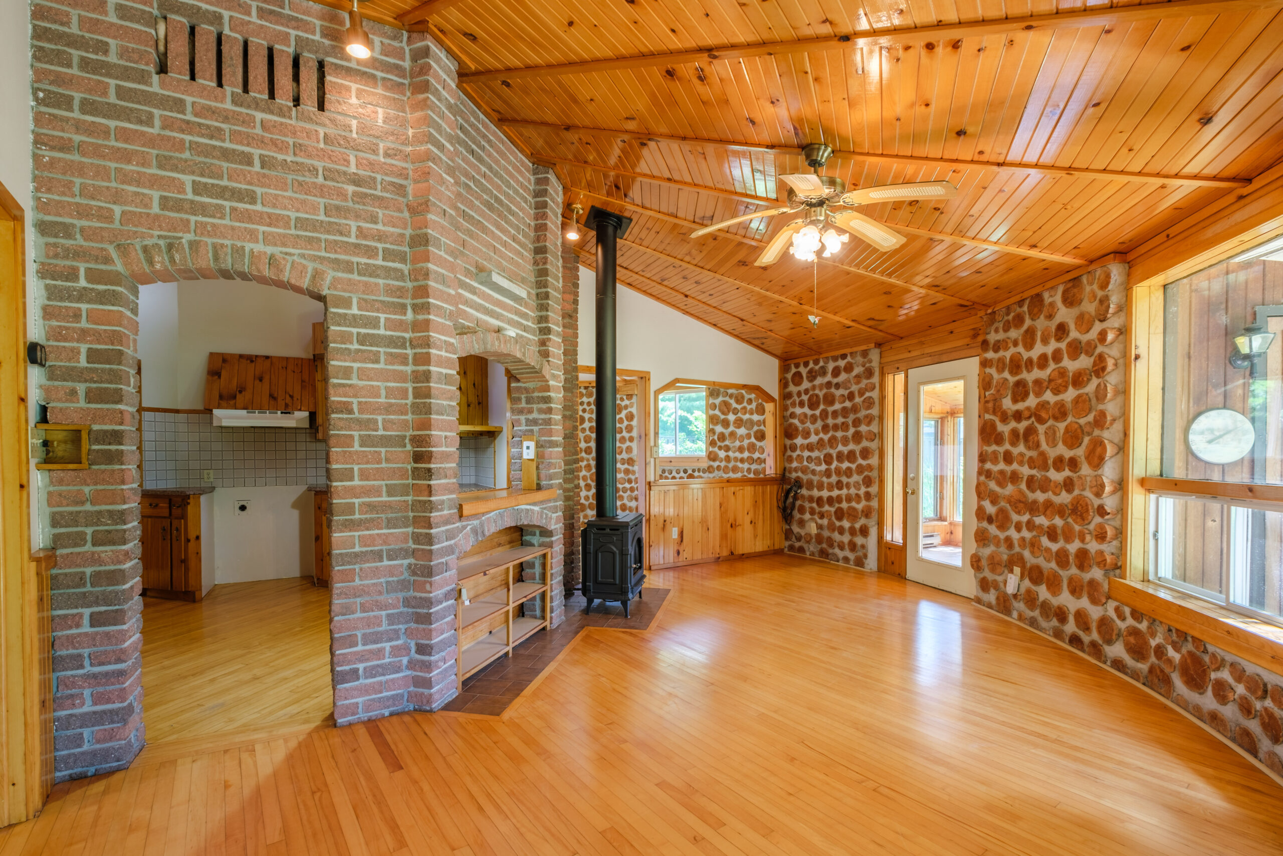 A brightly lit room with wood floors and ceiling. To the left, a stone-lined archway that leads into the kitchen, to the right a stone wall with large windows