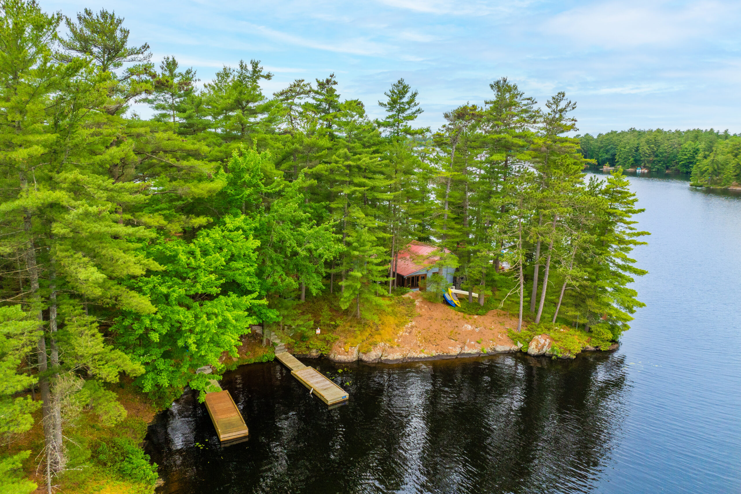 An island with tall trees has a small red cottage poking from the trees and two docks leading into the water