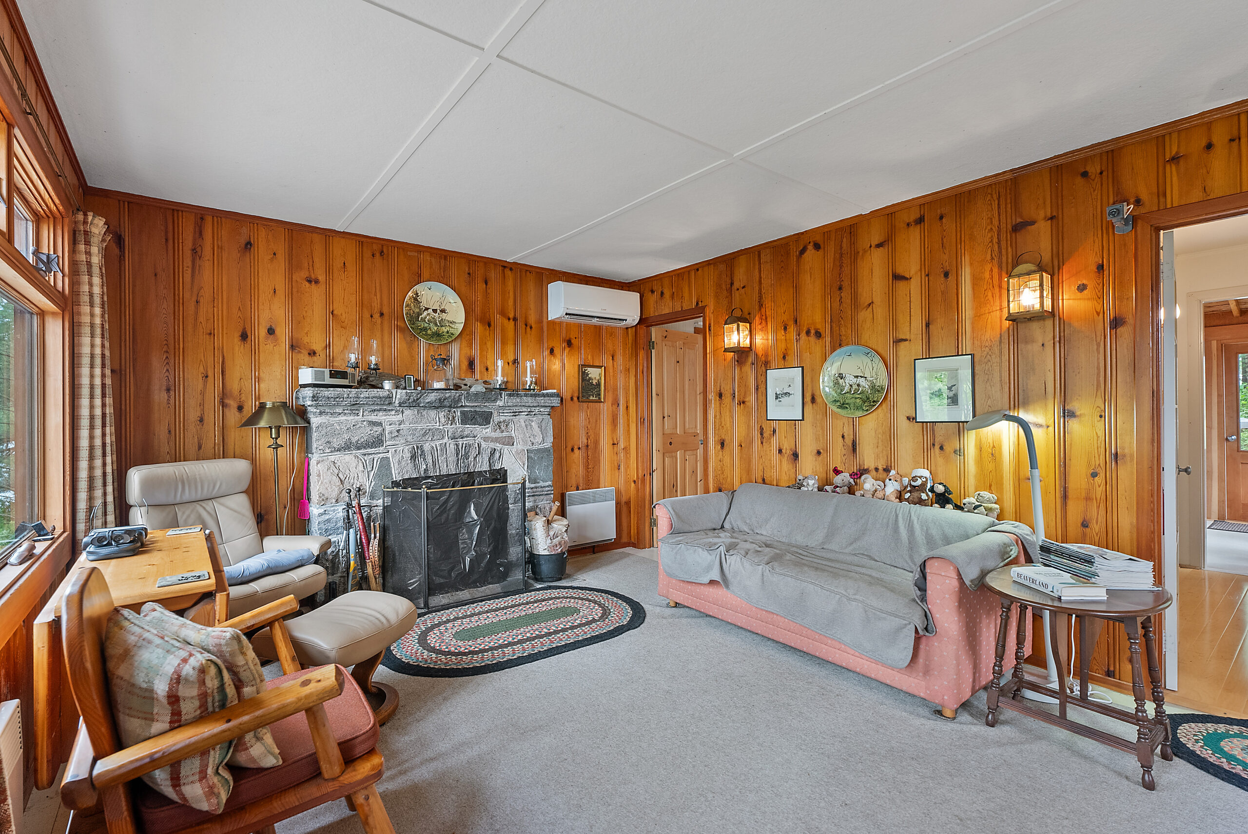A pink couch partially covered in a grey blanket faces a wood chair. In a wood room, a stone fireplace sits between them