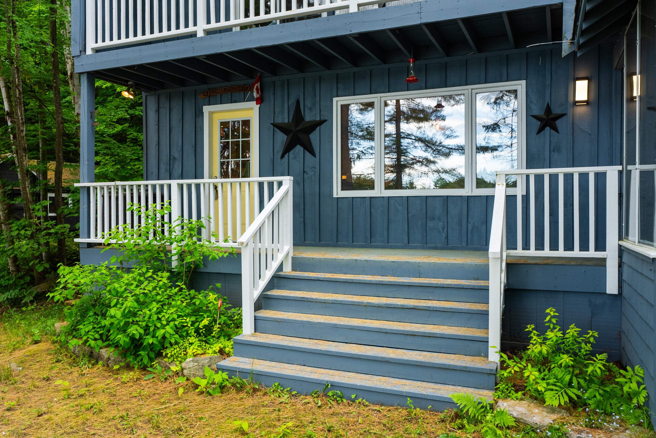 Steps with white railings lead up to a small balcony on the blue cottage. Bright windows are next to a yellow door