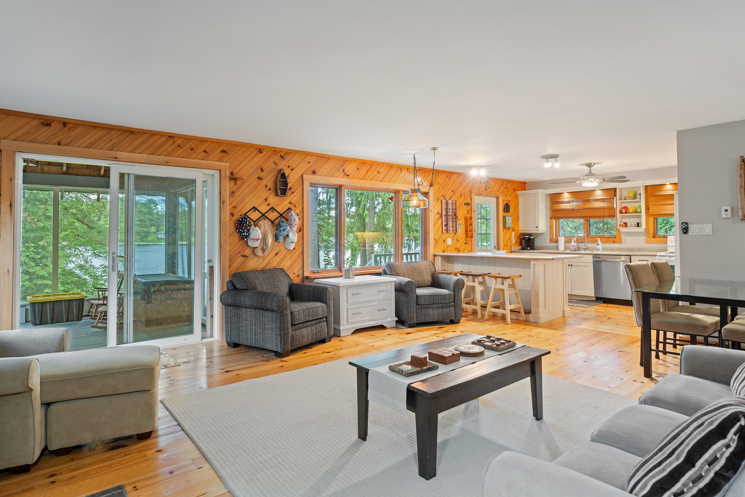 A coffee table on a white carpet in the centre of the room. On the far wall, two grey loveseats with a white table between. To the back-right, the kitchen