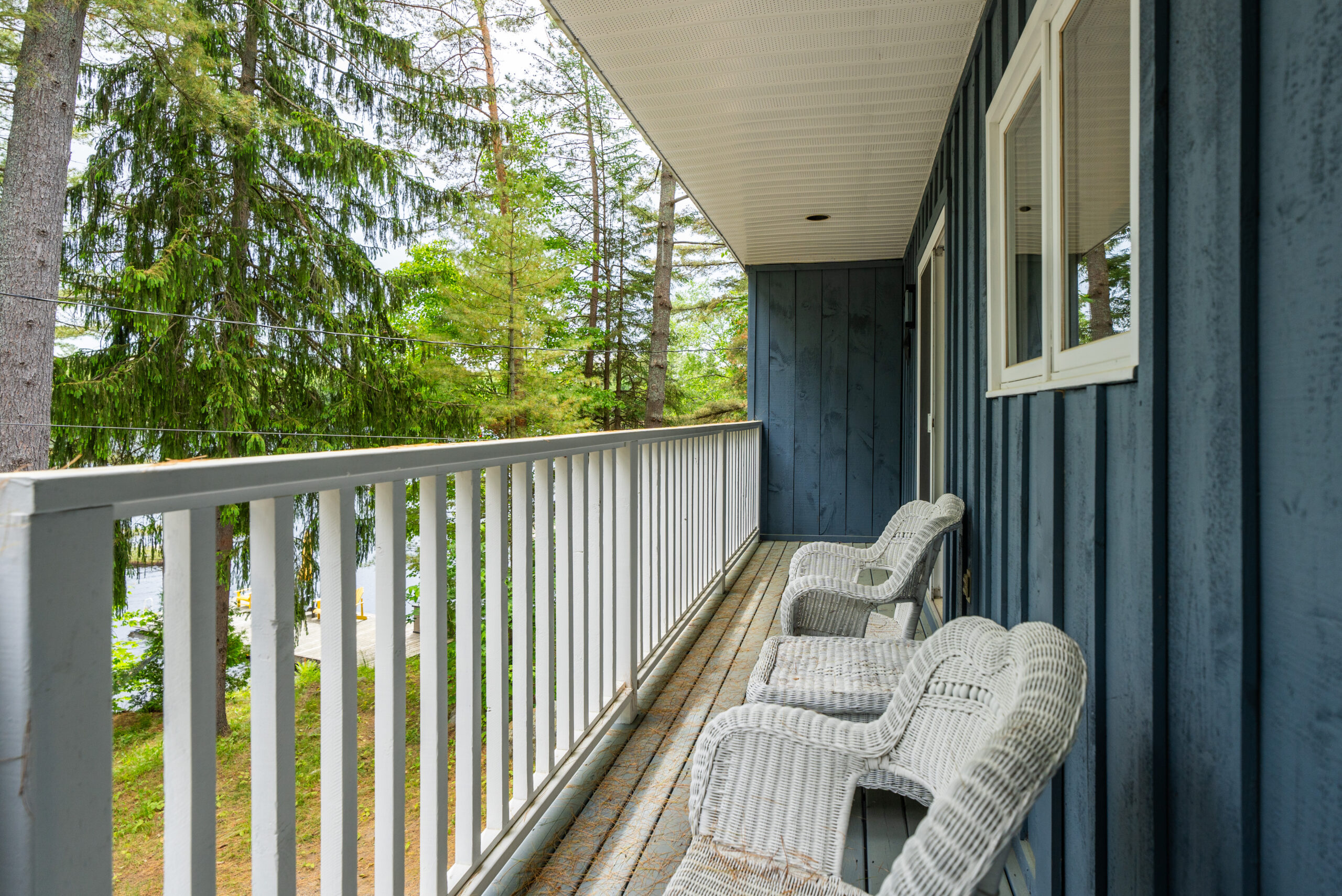 Two white wicker chairs sit against the blue panelled cottage. They face a white railing. Beyond the rail is trees with glimpses of the lake between