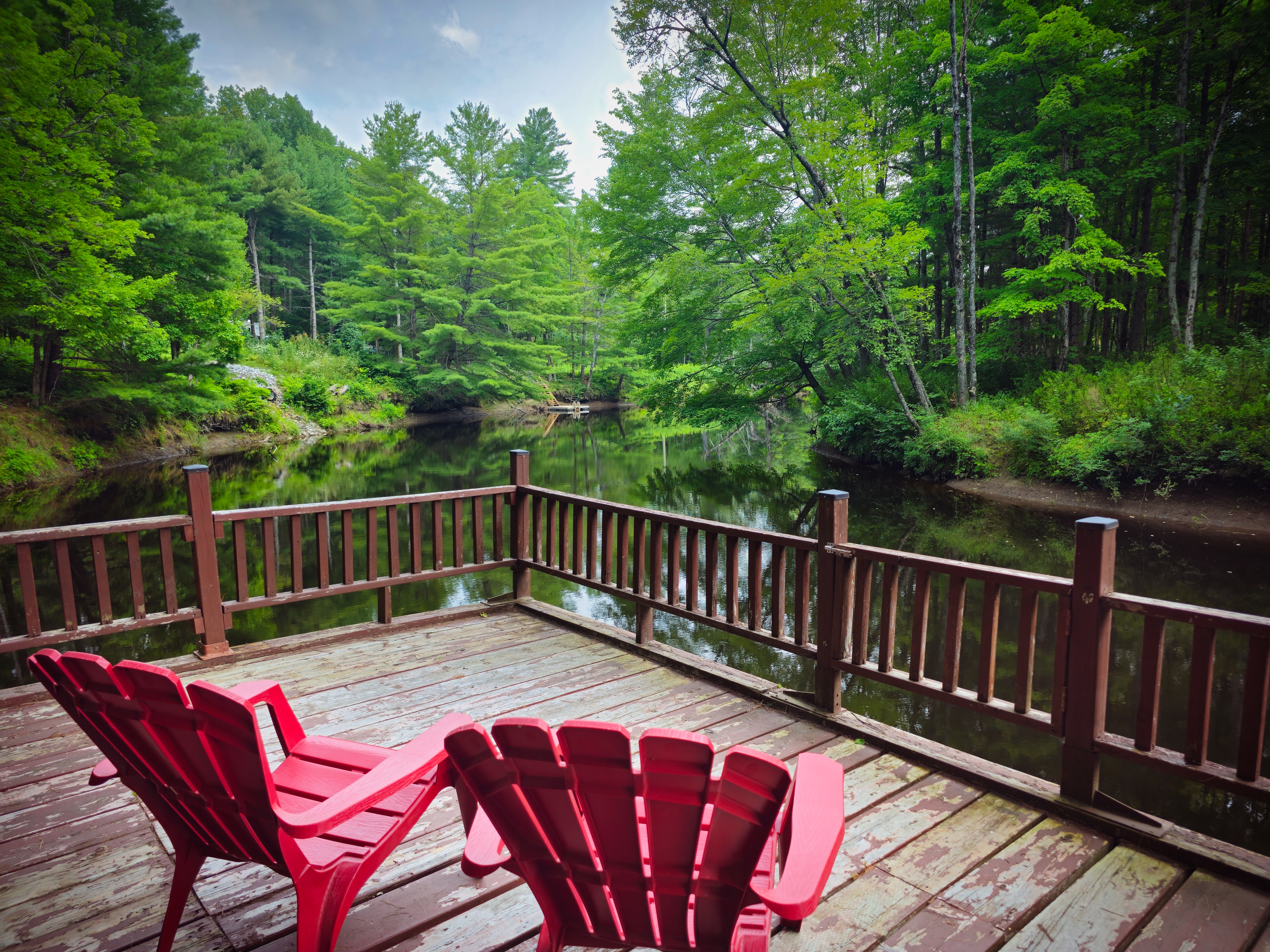 Two red Muskoka chairs on a wood deck face water