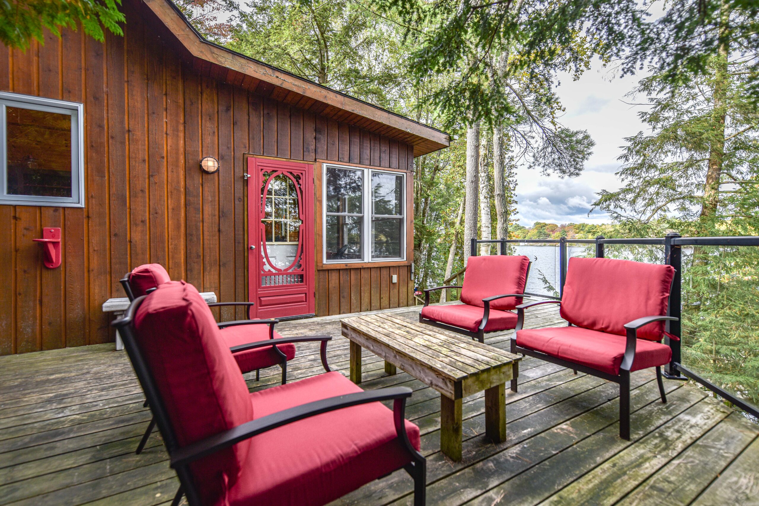 A set of four red chairs face a small wood table on the deck beside a wood cabin with a red door