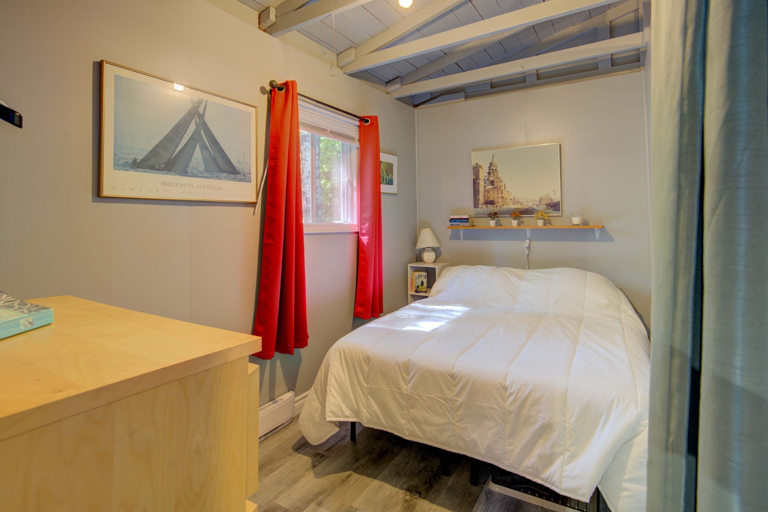 A large white bed beside a small window with red curtains