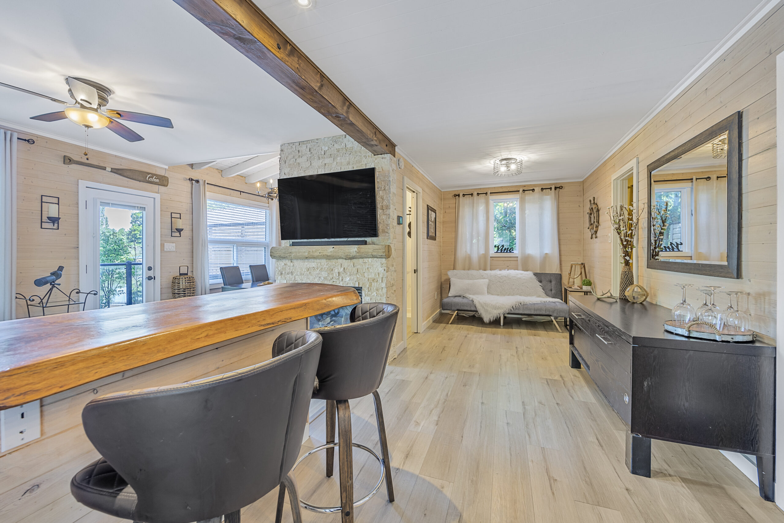 A bar table with stools. Behind, a flat screen TV hanging on a wall and a grey couch with a fluffy blanket
