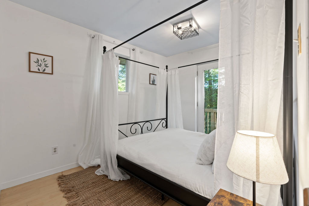 A white bed on a black bed frame with airy curtains. A door to the wraparound porch behind the bed
